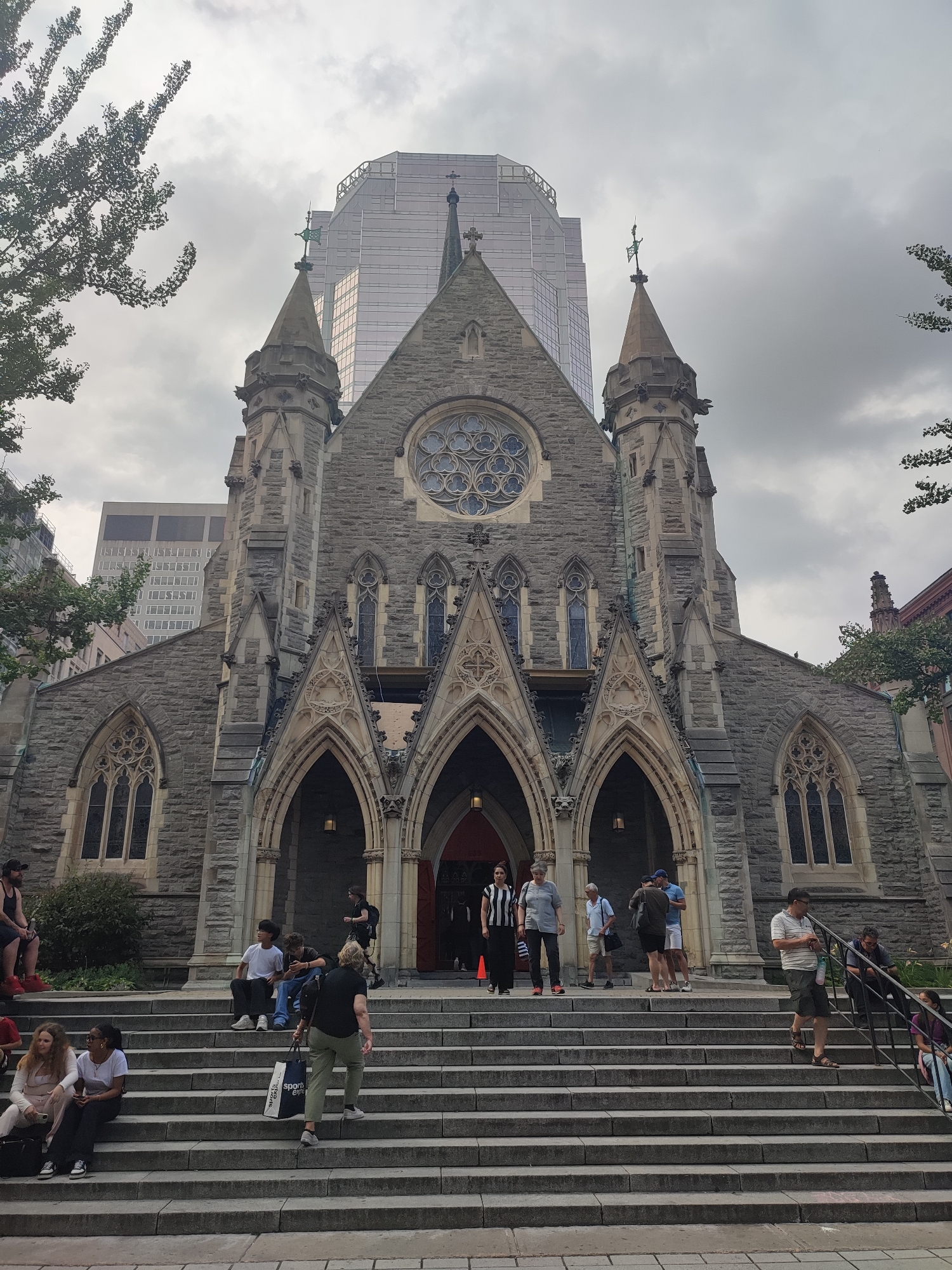 The image shows a stone church with gothic architectural features, including arched doorways, pointed windows, and spires. A modern glass skyscraper rises behind the church, suggesting an urban setting. Several people are visible on the wide stone steps leading up to the church entrance. Some are sitting, others are standing or walking, and one person is carrying a shopping bag. The sky is overcast, and trees are visible on the left side of the frame. This is the exterior of St. George's Anglican Church, located in Montreal, Canada.