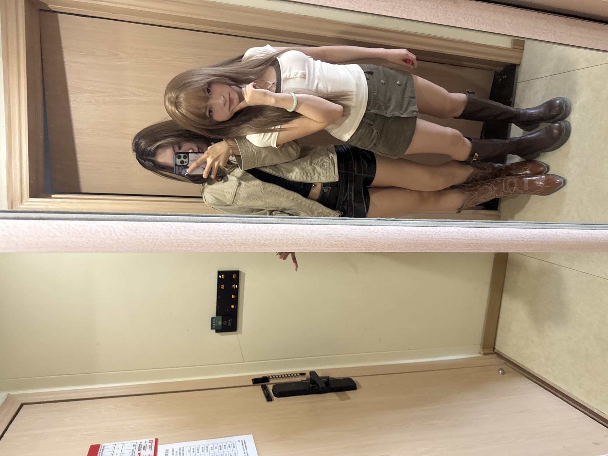 Two women are reflected in a mirror in an indoor setting. One woman is taking a photo of themselves and the other woman with a mobile phone. The woman in the foreground is wearing a tan jacket, a black top, a black skirt, and brown cowboy boots. The other woman is wearing a white top, a khaki skirt, and brown boots. The room has a beige wall, a wooden door, and a beige floor. A key card reader and a door lock are visible on the wall. A calendar is attached to the door. The location is likely in Dunhuang City, China.