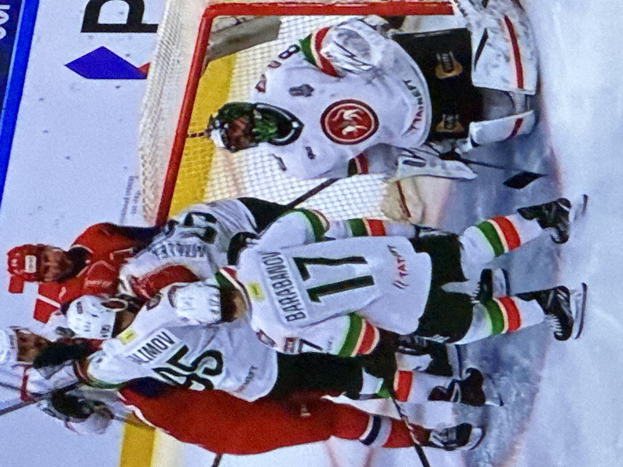 A photograph shows a hockey game being played in Kazan, Russia. The image is rotated approximately 90 degrees counterclockwise. Players wearing white jerseys with green and red stripes, identified by the names "BARABANOV" and "KLIMOV" on their backs, are involved in a scrimmage near the goal, guarded by a goaltender wearing a white mask and jersey adorned with a circular red and brown emblem. Opposing players in red jerseys are also part of the fray. The hockey net is visible in the background.