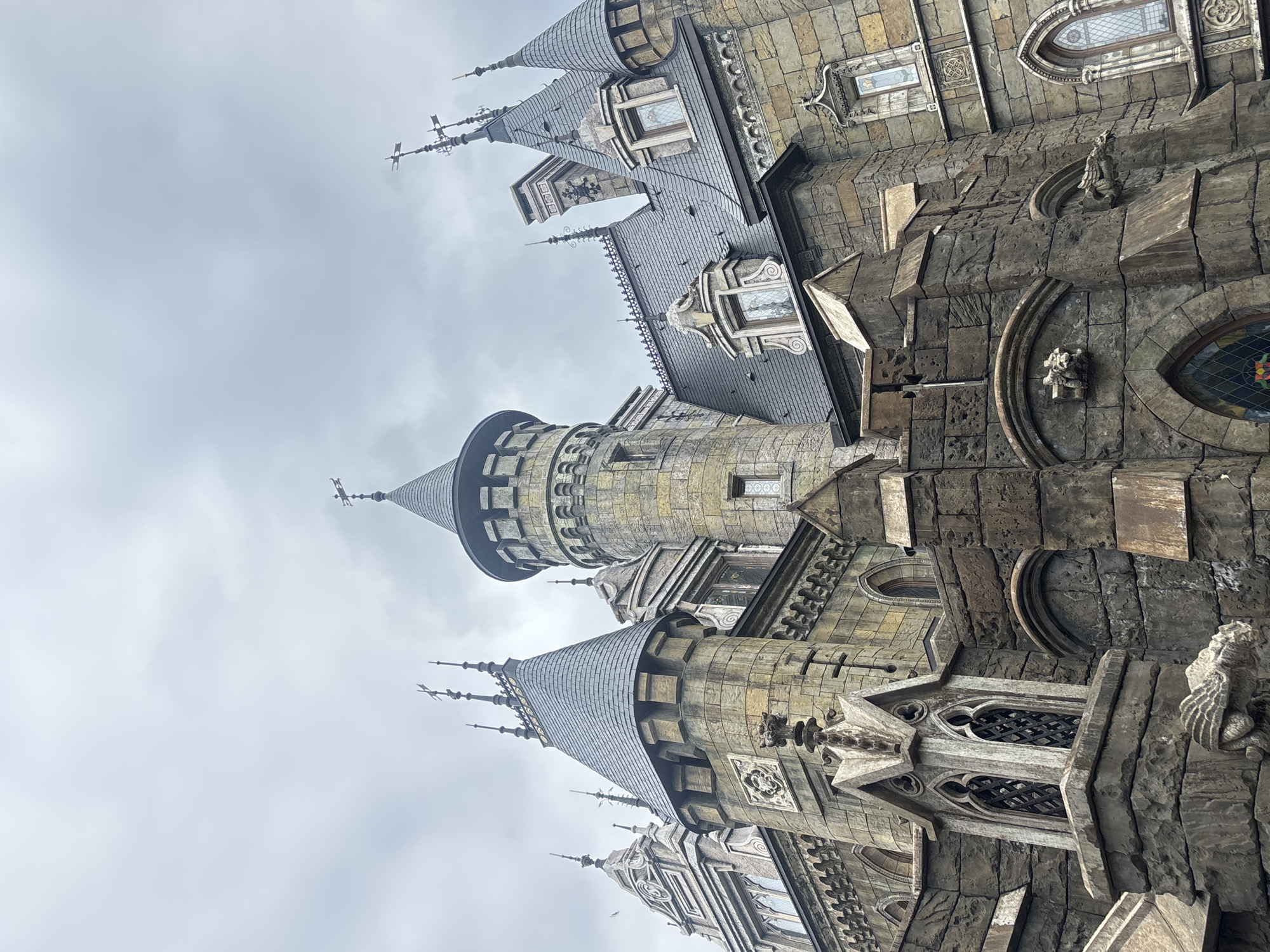 The image shows the exterior of the "Fantasysia Castle," a gothic-style architectural structure located in the city of Khryashchevka, Russia. The castle is made of stone and features multiple towers with conical roofs, pointed arches, and decorative gargoyles. The sky is overcast, suggesting a cloudy day. The perspective is from a low angle, looking up at the castle. No people are visible in the frame.