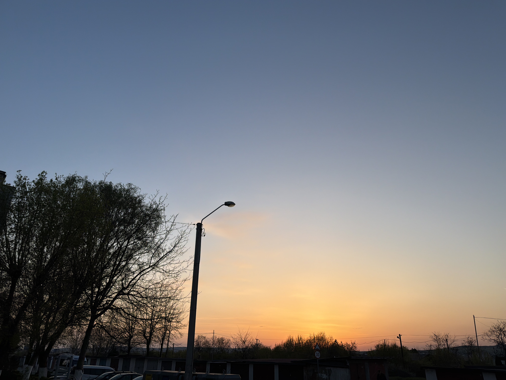 The image captures a sunset in what appears to be a peripheral area of Târgu Mureș, Romania. The sky transitions from a pale blue at the top to a warm orange near the horizon, suggesting the time of day. A street lamp stands prominently in the mid-ground, slightly offset from the center. To the left, there is a tree with dense branches, while on the ground, partially obscured by vegetation, are rows of what appear to be garage units and parked cars. The overall environment suggests an urban or suburban area, possibly an industrial zone or the edge of a residential neighborhood. The scene is static, with no apparent actions or interactions visible.
