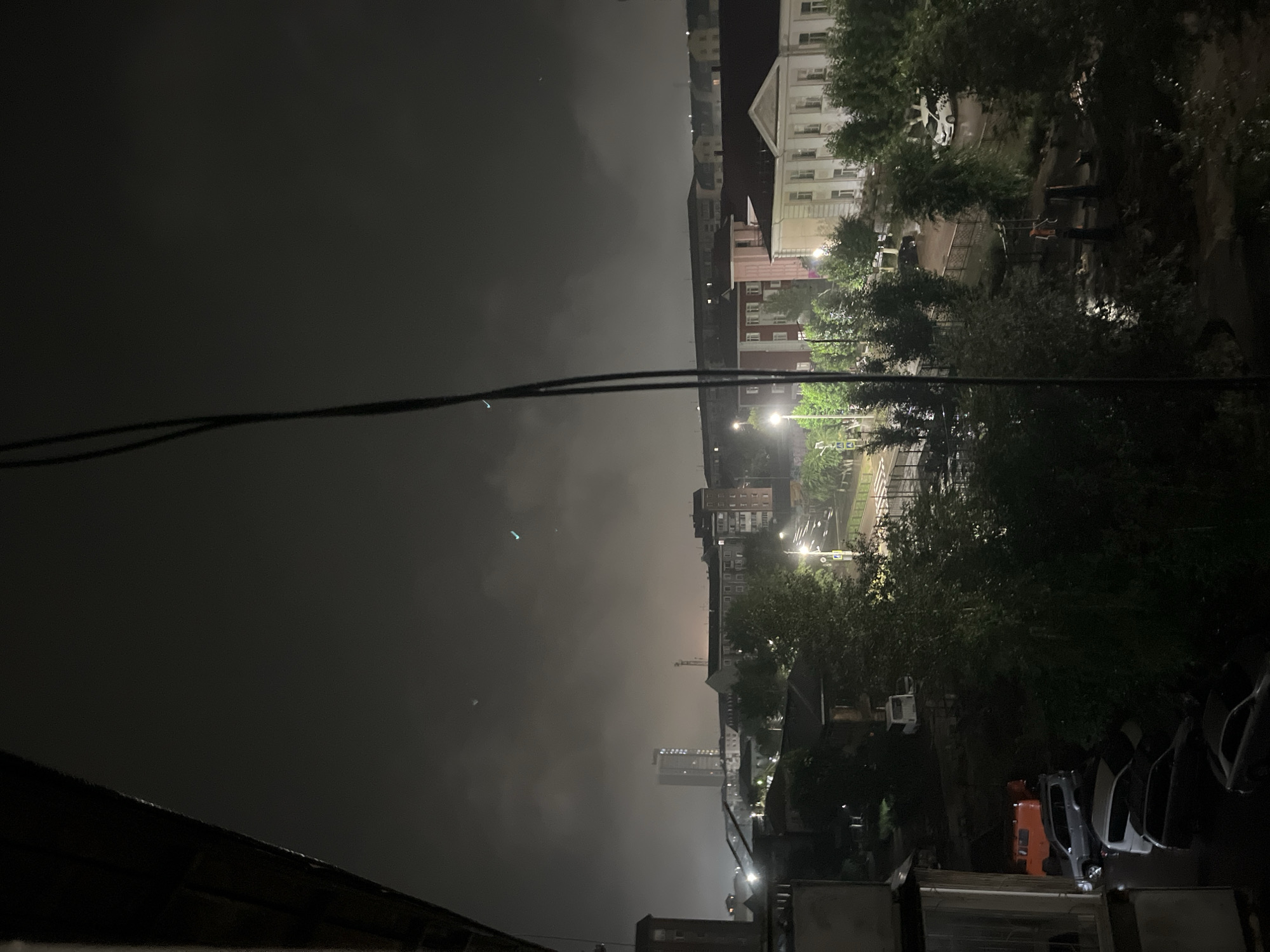 A nighttime exterior shot captures a hazy, overcast sky above a residential area. In the distance, a tall, illuminated building stands out against the darkness, flanked by other multi-story buildings. A street with illuminated lights and passing car lights is visible, suggesting moderate traffic. Closer to the foreground, several cars are parked on a street or in a lot, some partially obscured by trees. The overall scene depicts a somewhat urban environment under a low-light, possibly humid or foggy, atmospheric condition.