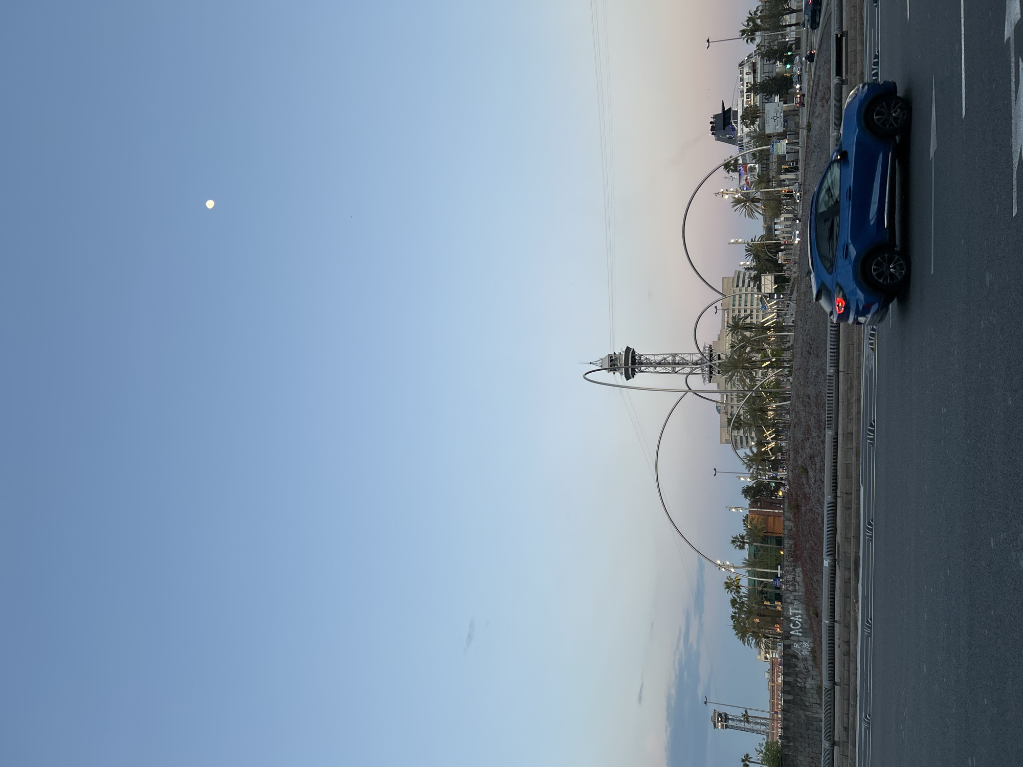 A slightly rotated, horizontal wide shot captures an urban twilight scene in what appears to be Barcelona, Spain. The sky dominates the upper left, transitioning from a soft blue to a faint, warm pinkish hue on the right, indicative of either dusk or dawn. A distinct, bright white moon is visible in the upper left quadrant of the sky.
In the foreground, a paved road stretches along the right side of the frame, marked with a white arrow indicating forward movement. A modern, dark blue car, likely an SUV or crossover, is positioned on the right lane, facing away from the viewer with its taillights faintly glowing. To the left of the road, a concrete barrier separates it from a cultivated area featuring small shrubs and palm trees.
The middle ground and background are characterized by urban infrastructure. Numerous palm trees are scattered among buildings. A prominent, distinctive tower-like structure with large, curving metallic arches or rings stands centrally, possibly part of an observation deck or cable car system. Other buildings, some with illuminated windows or streetlights, line the horizon.
Visible text includes a white arrow painted on the road and graffiti spelling "ACAT" on a low wall or barrier near the road in the mid-ground. The overall mood is calm, capturing a transition hour in a bustling city environment.