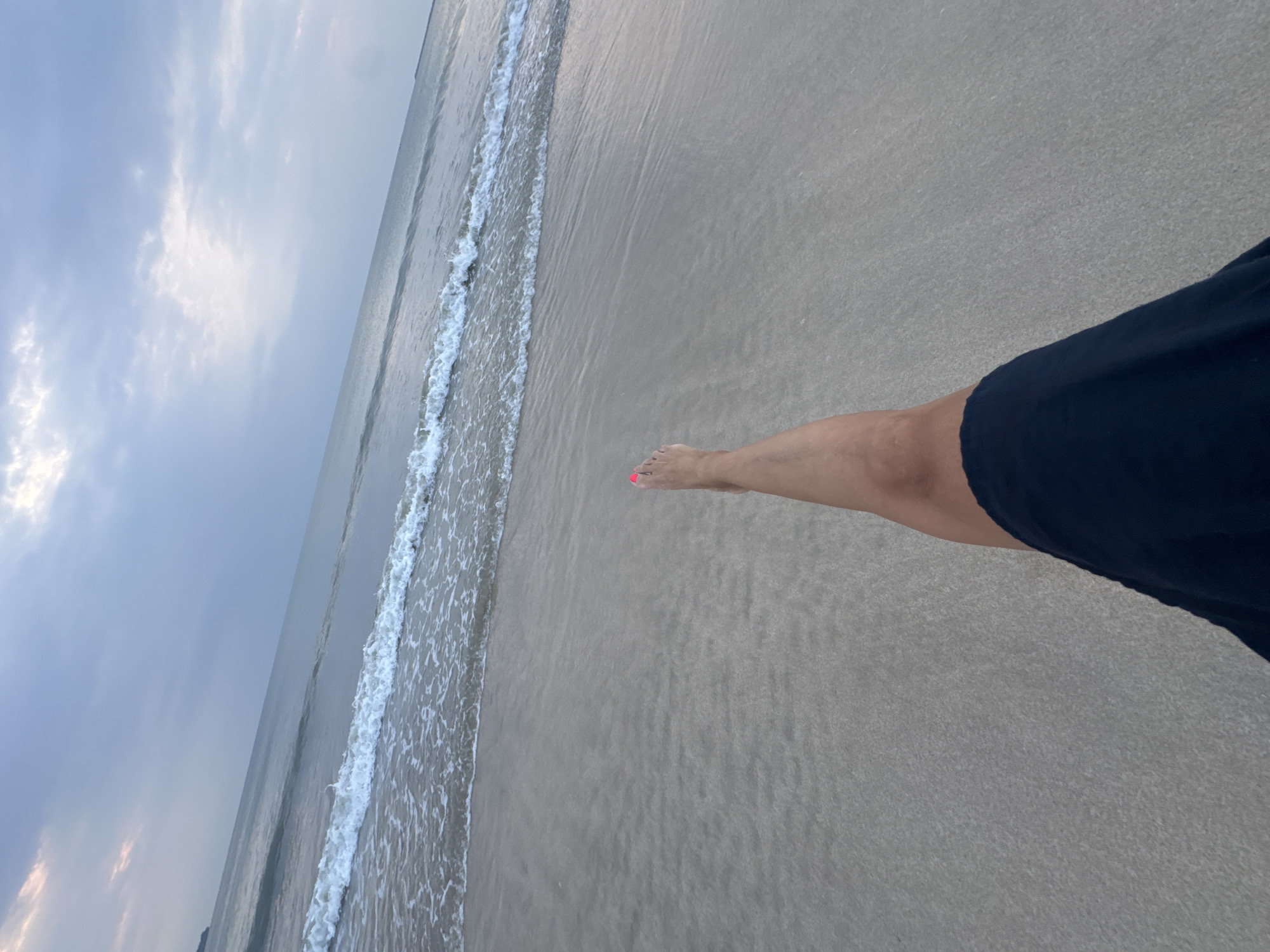 The image depicts a first-person perspective on a sandy beach in Jimbaran, Indonesia. A bare left leg is extended towards the ocean, with the big toenail exhibiting bright pink polish. The lower right foreground reveals a portion of the subject's right thigh and knee, covered by dark blue or black fabric. The immediate environment consists of wet, light-colored sand, merging with shallow ocean water where small white waves are breaking. The open sea extends to the horizon beneath an overcast sky characterized by light blue-grey cloud formations. A faint landmass is discernible on the distant left horizon.