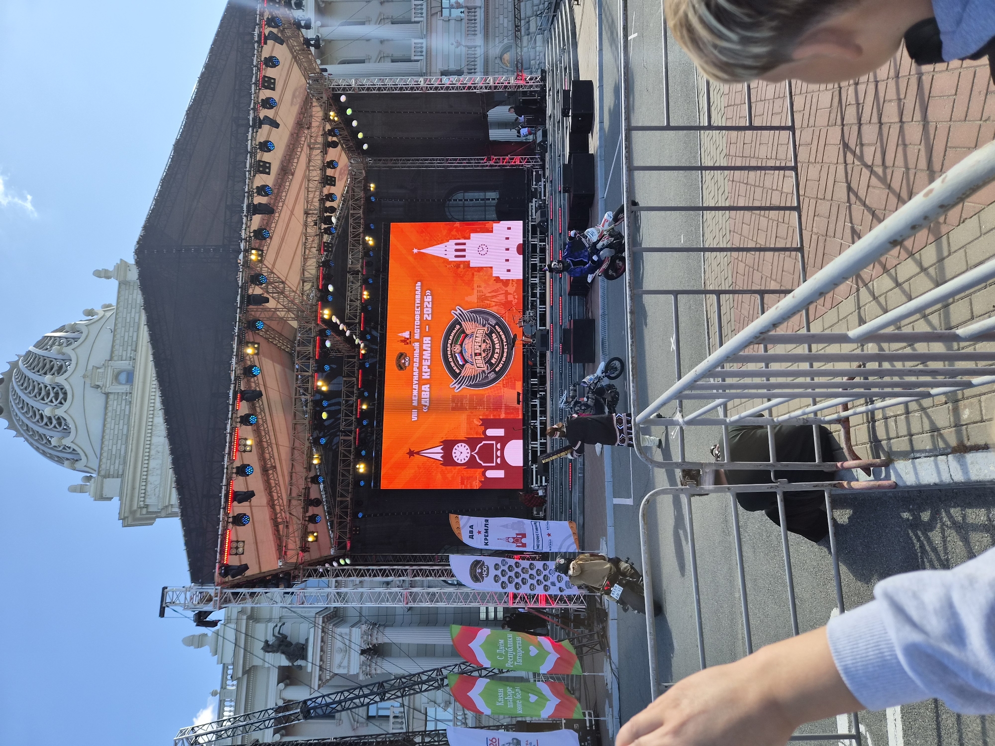 The image depicts an outdoor event in Kazan, Russia, likely a festival or concert, set against the backdrop of a grand, white building with a dome, reminiscent of a palace or government structure. In the foreground, a temporary stage with a large LED screen is erected. The screen displays the text "VIII МЕЖДУНАРОДНЫЙ МОТОФЕСТИВАЛЬ «ДВА КРЕМЛЯ - 2026»" (VIII INTERNATIONAL MOTORCYCLE FESTIVAL "TWO KREMLINS - 2026"), along with stylized images of the Kazan Kremlin and a motorcyclist emblem.
Several people are present: a person in traditional attire stands with a long object, possibly a staff, while two individuals on motorcycles are visible in the mid-ground, one moving and another stationary. Spectators are seen behind a metal barrier, with a child's head in the lower right corner looking towards the event. Flags with Russian and Tatar inscriptions are positioned to the left of the stage. The scene is illuminated by daylight, with a clear blue sky overhead. The overall environment suggests a public gathering in a prominent urban setting.