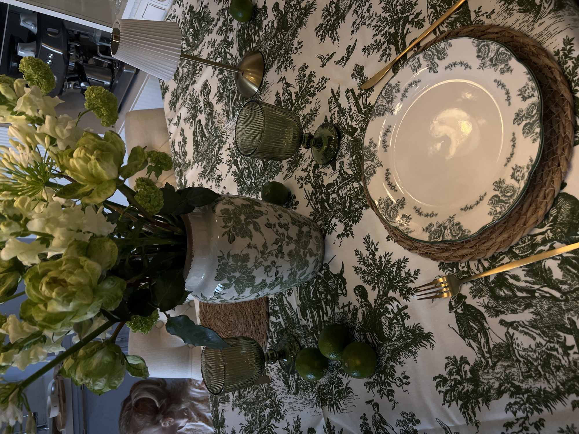 The image captures an elegantly set dining table in what appears to be an indoor setting in Moscow, Russia. The table is covered with a white tablecloth featuring an intricate green toile de Jouy pattern, depicting various pastoral scenes with figures, animals like donkeys, and lush trees.
A large, off-white ceramic vase, adorned with a matching green floral pattern, holds a vibrant bouquet of fresh flowers. The bouquet predominantly features white and light green blooms, including what resemble full-petaled roses or hydrangeas and spiky white flowers.
A single place setting is visible, comprising a round, woven straw placemat. On top, a white ceramic dinner plate with a scalloped edge is decorated with a delicate green floral border. Gold-colored cutlery, a fork and a knife, are placed beside the plate. Scattered across the tablecloth are several bright green limes, adding a fresh accent. Two dark green, ribbed glass goblets or tumblers are present; one stands upright, while the other lies on its side.
In the upper background, a small table lamp with a pleated white shade and a gold stem casts soft light, contributing to the intimate ambiance. Reflections on the plate and glassware suggest warm, artificial illumination. The blurry background offers glimpses of kitchen appliances, possibly a coffee machine, and an upholstered cream-colored chair or sofa. A person's head with light, curly hair is partially visible and out of focus below the table, suggesting active participation in setting or enjoying the space.
The scene conveys the preparation for a refined meal or special gathering. The overall color scheme is dominated by whites and various shades of green, accented with gold, creating a fresh yet classic aesthetic. No visible text is present in the image.