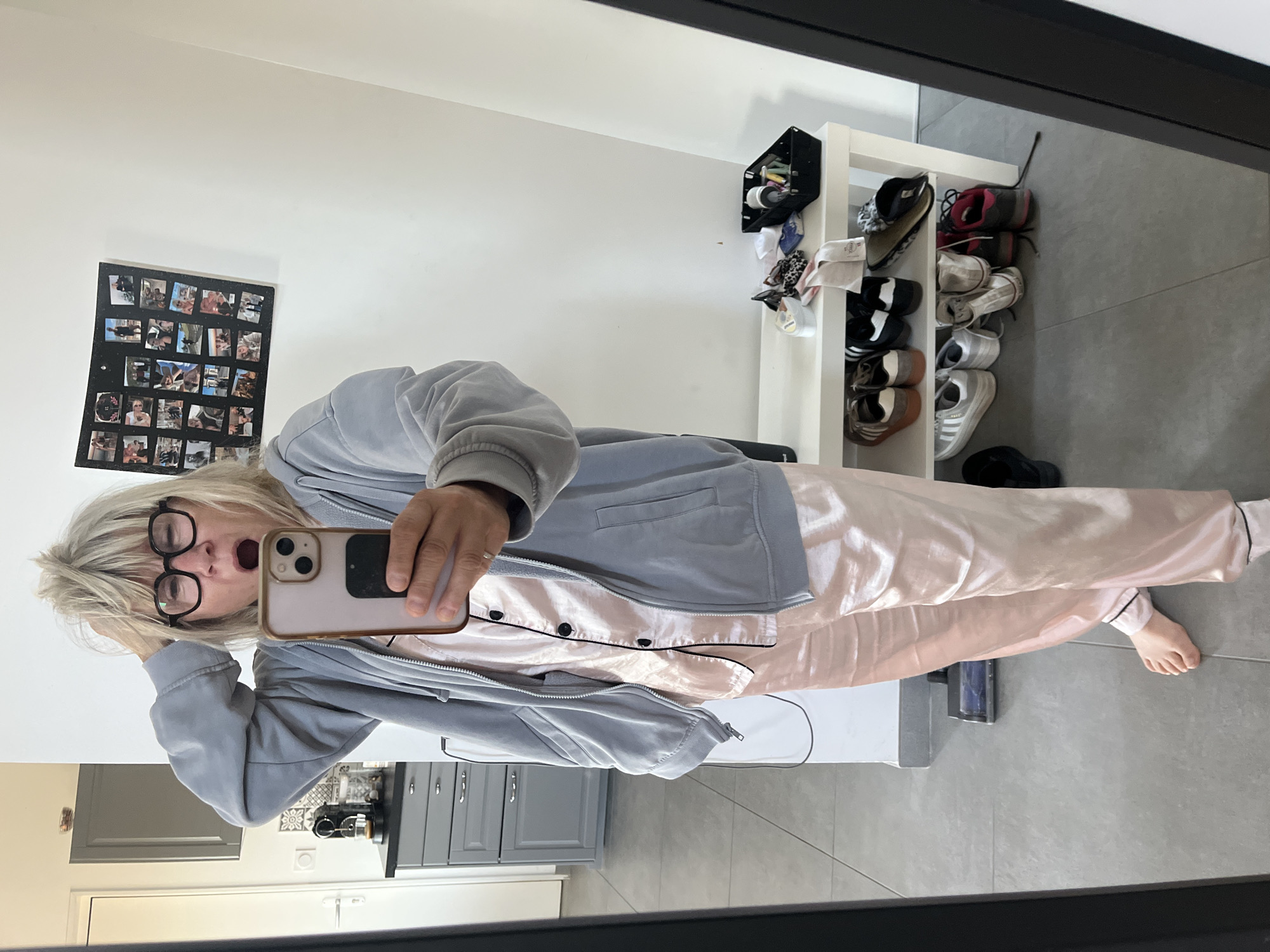 A woman is taking a selfie in a room, likely her home, in Chambœuf, France. She is wearing a gray hooded sweatshirt over pink silk pajamas. She has blonde hair and is wearing glasses. She is holding a smartphone in her right hand and has her left hand on her head, with her mouth open as if yawning or speaking. The room appears to be a domestic interior, possibly a bedroom or living area, with a mirror that is reflecting the scene. In the background, a shoe rack filled with shoes, a small shelf with various items, and a wall decorated with a collage of photographs are visible. The floor is tiled.
