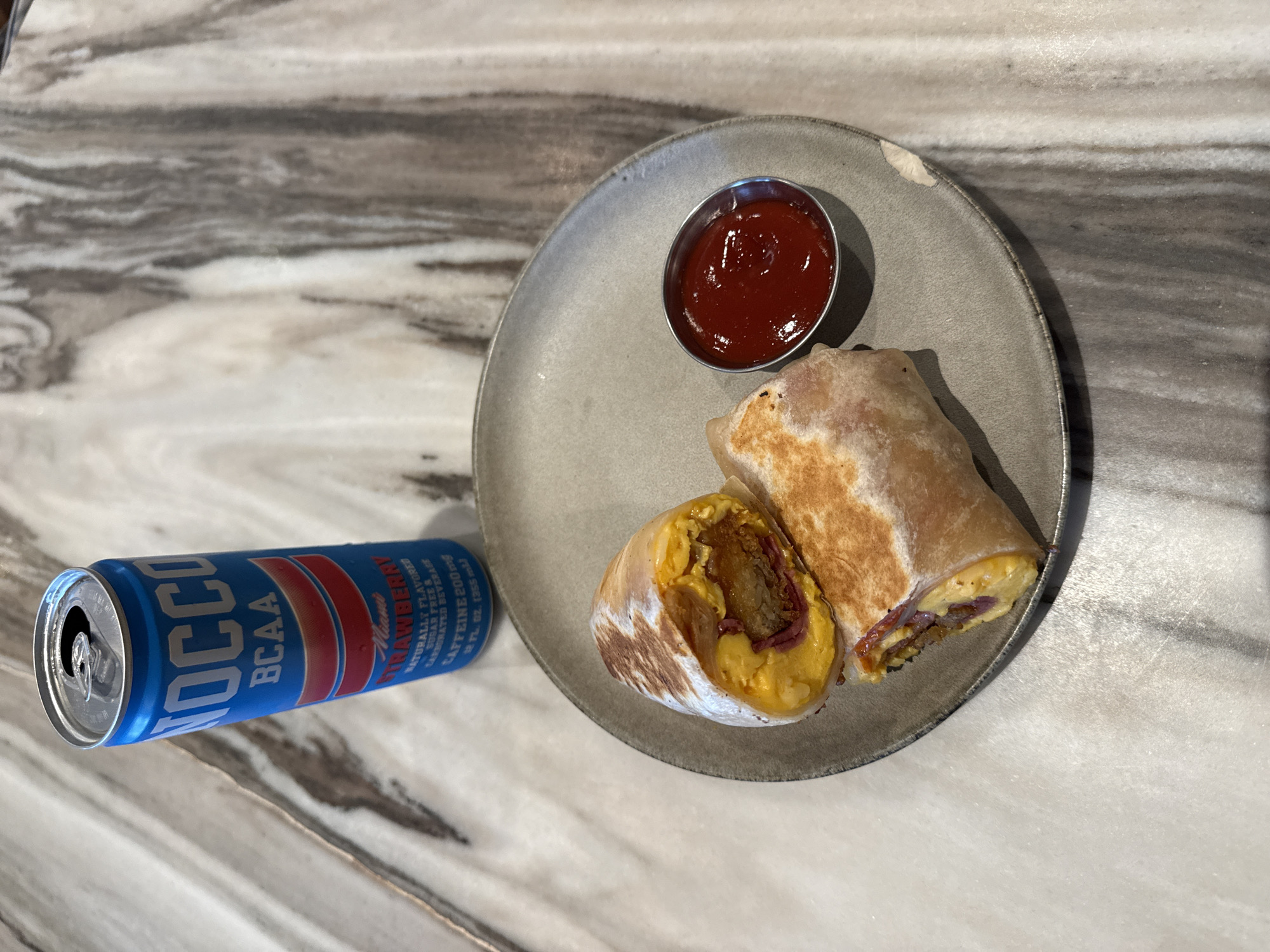 The scene features a plate of food and a can of a beverage on a marbled countertop.
On the plate, there are two pieces of what appears to be a breakfast burrito, served with a small dish of ketchup or red sauce. The burrito seems to contain eggs and some type of meat.
Next to the plate, resting on the countertop, is a blue can of "NOCCO BCAA Miami Strawberry" flavored energy drink. The countertop itself is a marbled pattern in shades of white, gray, and brown.
The location is likely a kitchen or dining area with a marbled countertop, situated in Los Angeles, United States of America.