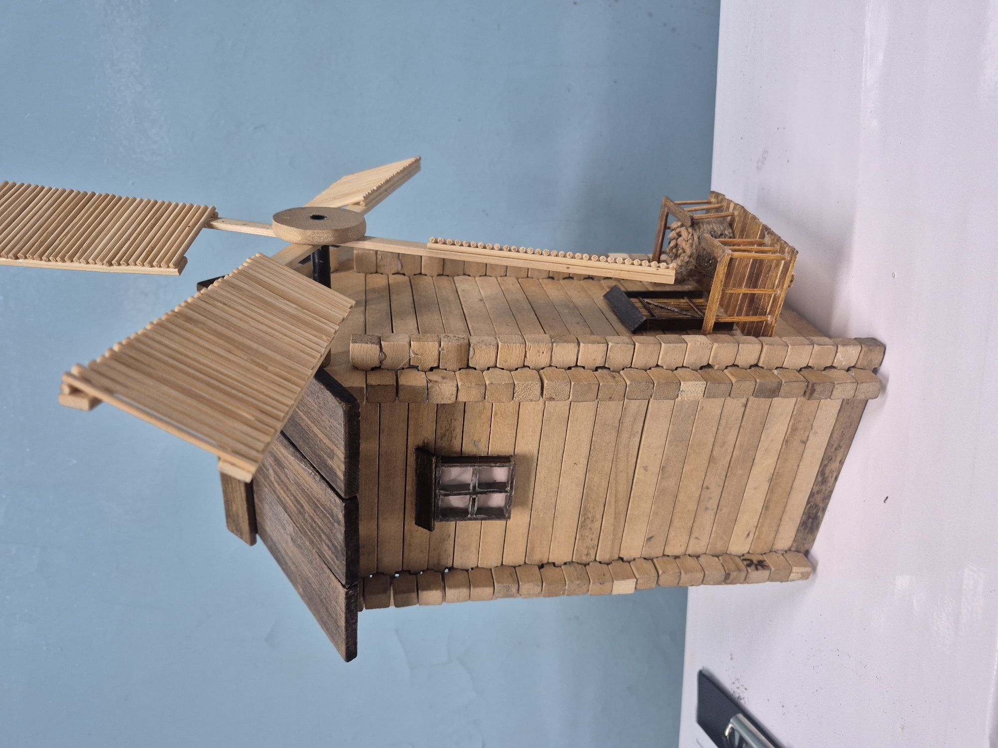 A craft project depicting a windmill is positioned against a two-tone backdrop of pale blue and white. The windmill is constructed primarily of popsicle sticks. It features four blades made of popsicle sticks arranged in a fan-like structure. The body of the windmill is also composed of popsicle sticks, arranged horizontally and vertically to create the walls and roof. A small, dark-framed window is present on one side of the structure. On the roof, a miniature wooden structure resembling a hayloft, complete with a miniature pile of straw, is visible. The edges of the windmill’s structure are accented with popsicle sticks placed end-to-end, creating a textured border. The overall color of the windmill is a light tan, with some elements, such as the front door, stained a darker brown. The setting is indoors, as evidenced by the presence of the white background, which appears to be a wall or table surface. The object appears to be rotated to the side for the purpose of the photo. The location information cannot be verified based on the image.