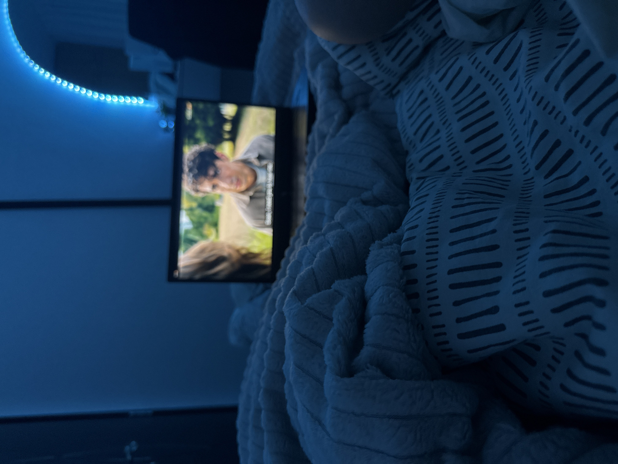 The image shows a person in bed with a laptop displaying a movie or show. The bed has a soft, textured white blanket and a pillow with a linear pattern. The room is dimly lit with a blue hue, possibly from LED lights on the wall. There is furniture visible in the background. The location in Blagoevgrad, Bulgaria cannot be determined from the visual information in the image.