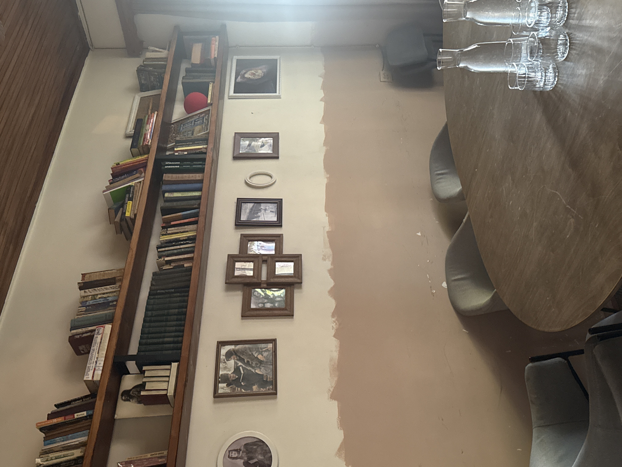 A view of a room with a bookshelf and a table with chairs in Batumi, Georgia. The bookshelf is filled with books and some other objects. There are framed pictures on the wall next to the bookshelf. The table has water bottles and glasses on it, and there are chairs around the table. The walls are painted in two different colors, and one part looks unfinished.