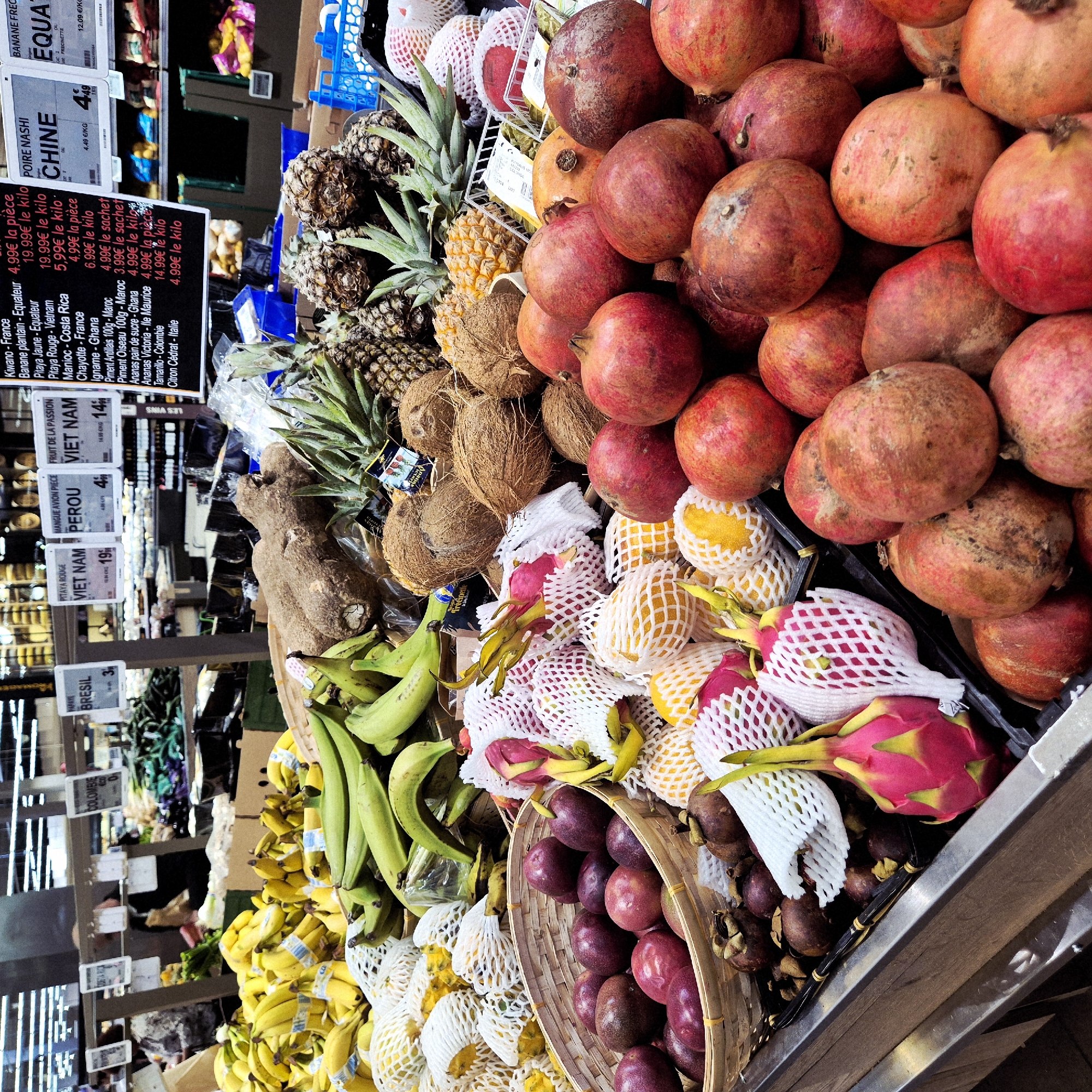 The image shows a display of fruits at a grocery store. Fruits visible include pomegranates, coconuts, pineapples, bananas, plantains, dragon fruit, and passion fruit. Price tags are visible. Text on signs includes "Kiwano-France", "Banane plantain-Equateur", "Pitaya Jaune-Equateur", "Manioc-Costa Rica", "Chayotte-France", "Igname-Ghana", "Piment Antillais 100g-Maroc", "Piment Oiseau 100g-Maroc", "Ananas pain de sucre-Ghana", "Ananas Victoria-Ile Maurice", "Tamarillo-Colombie", "Citron Cedrat-Italie", "Pitaya Rouge", "Mango Avion Piece", "Fruit de la Passion", "Poire Nashi Chine", and "Banane Frecinette Equateur". Text on the signs include prices in Euros. The location is Geispolsheim, France.