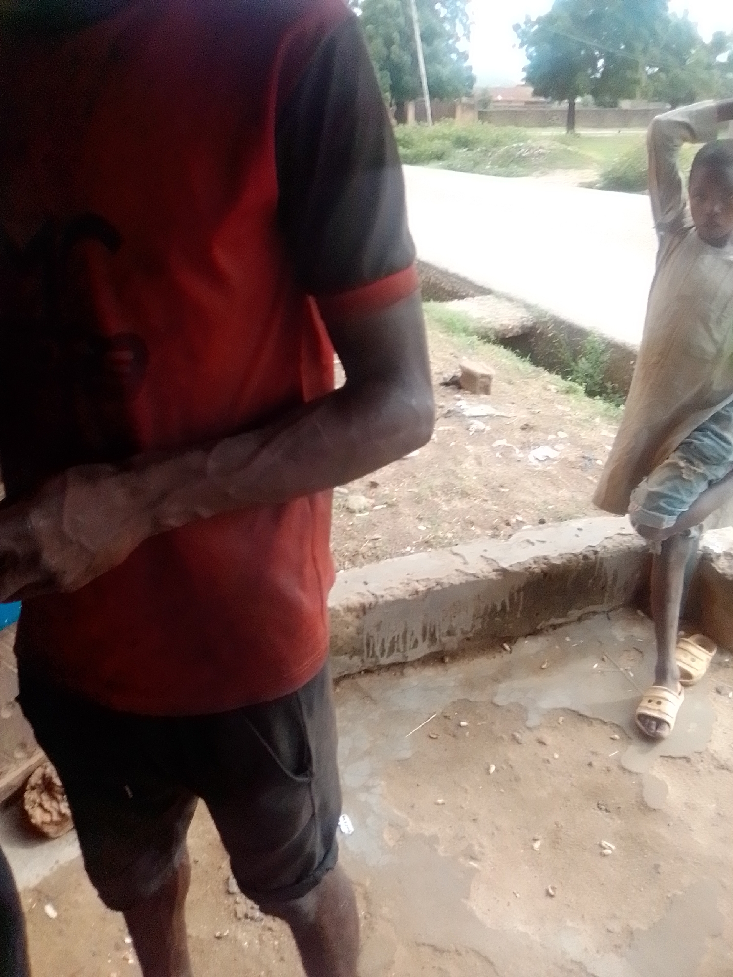 The image shows two individuals in an outdoor setting, possibly Kwadon, Nigeria. On the left, a person is wearing a red t-shirt with black sleeves and black shorts. Their left arm is visible, showing prominent veins. On the right, a second person is wearing a light-colored, long-sleeved garment and what appear to be light-colored sandals. The ground is uneven and appears to be dirt and concrete, with some scattered debris. A low concrete structure runs along the middle ground. In the background, trees and some buildings are visible, suggesting a semi-urban or rural environment. The lighting suggests it is daytime.