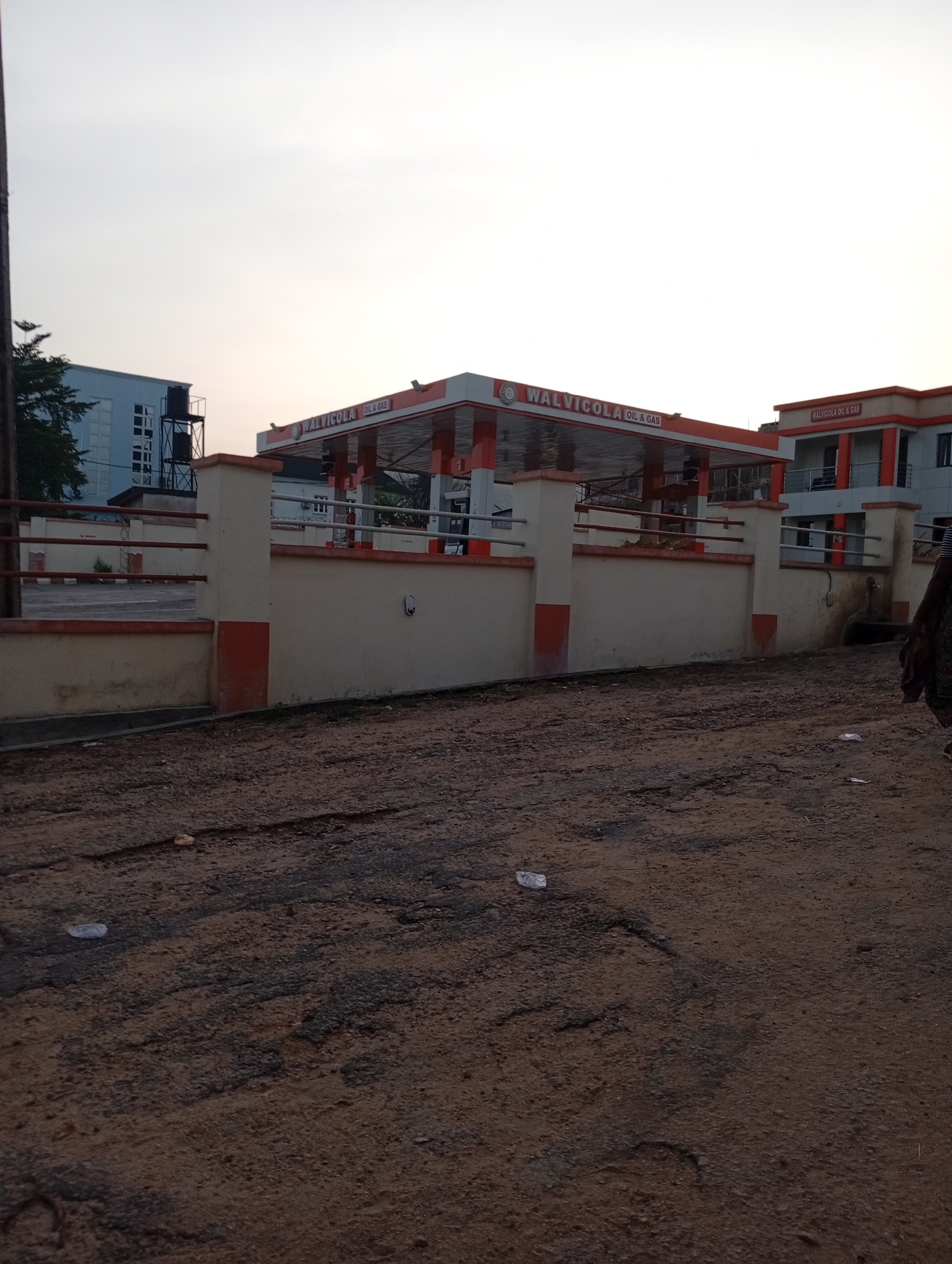 The image depicts a daytime scene in Adankolo, Nigeria, featuring a "WALVICOLA OIL & GAS" station. The station has a covered fueling area and an adjacent two-story building with similar branding. A low wall with pillars and railings surrounds part of the station. The foreground shows a rough, unpaved street with scattered debris. A partial view of a person is visible on the right side of the image. A taller building with a water tower is visible behind the gas station. The sky is overcast.