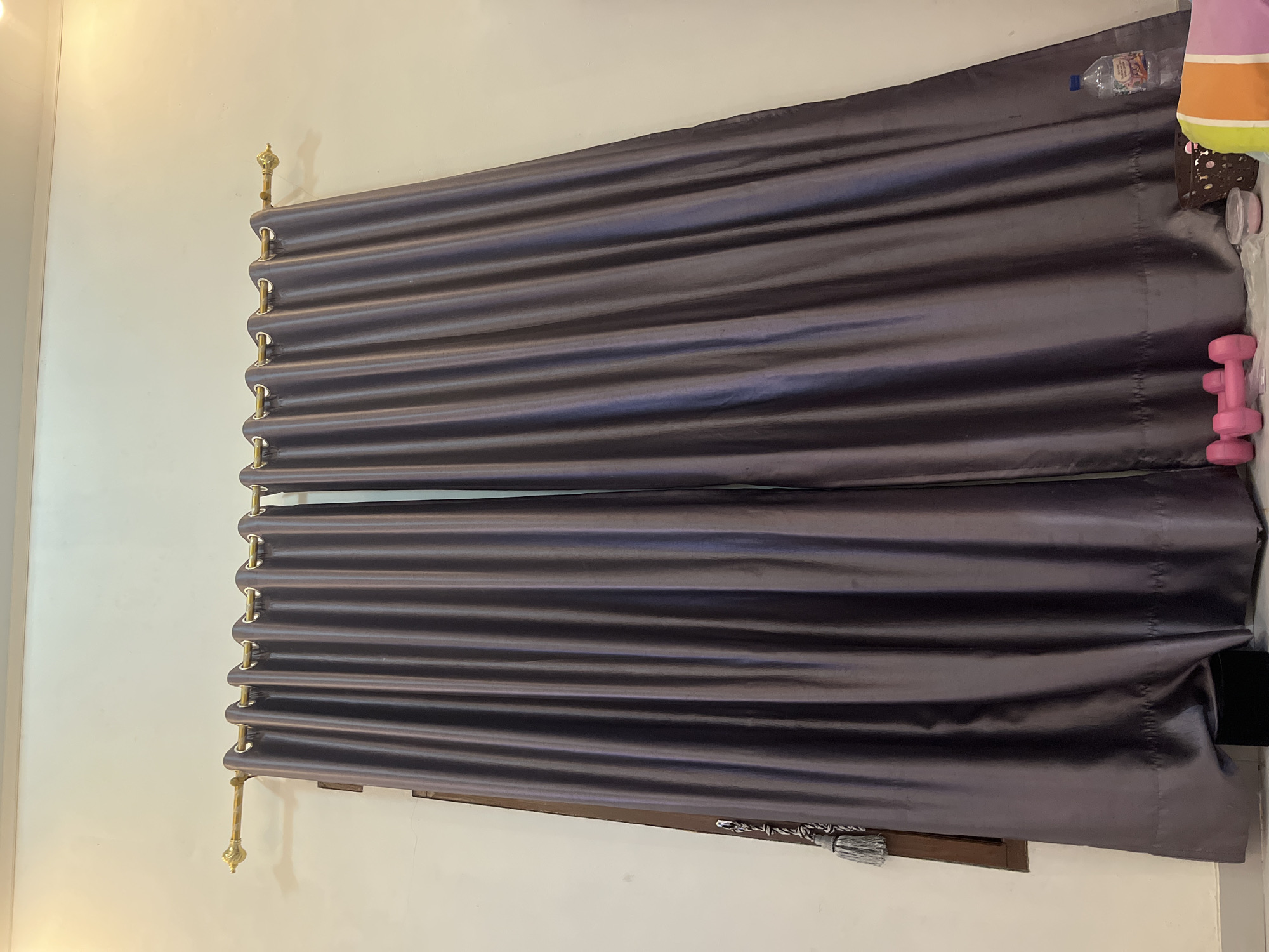The image depicts a window covered by two dark grey curtains, suspended from a gold curtain rod. The curtains are drawn closed, obscuring the view outside. On the left side of the image, a dark object, possibly a curtain tie-back with a tassel, is visible near the window frame. The lower portion of the image shows a black rectangular object on the floor, likely a base or stand. To the right, a pink dumbbell and a clear plastic water bottle are present. The background is a plain, off-white wall. The scene is indoors, with the lighting suggesting artificial illumination. There are no people visible in the image. Based on the information provided, this scene is located in Kebumen, Indonesia.