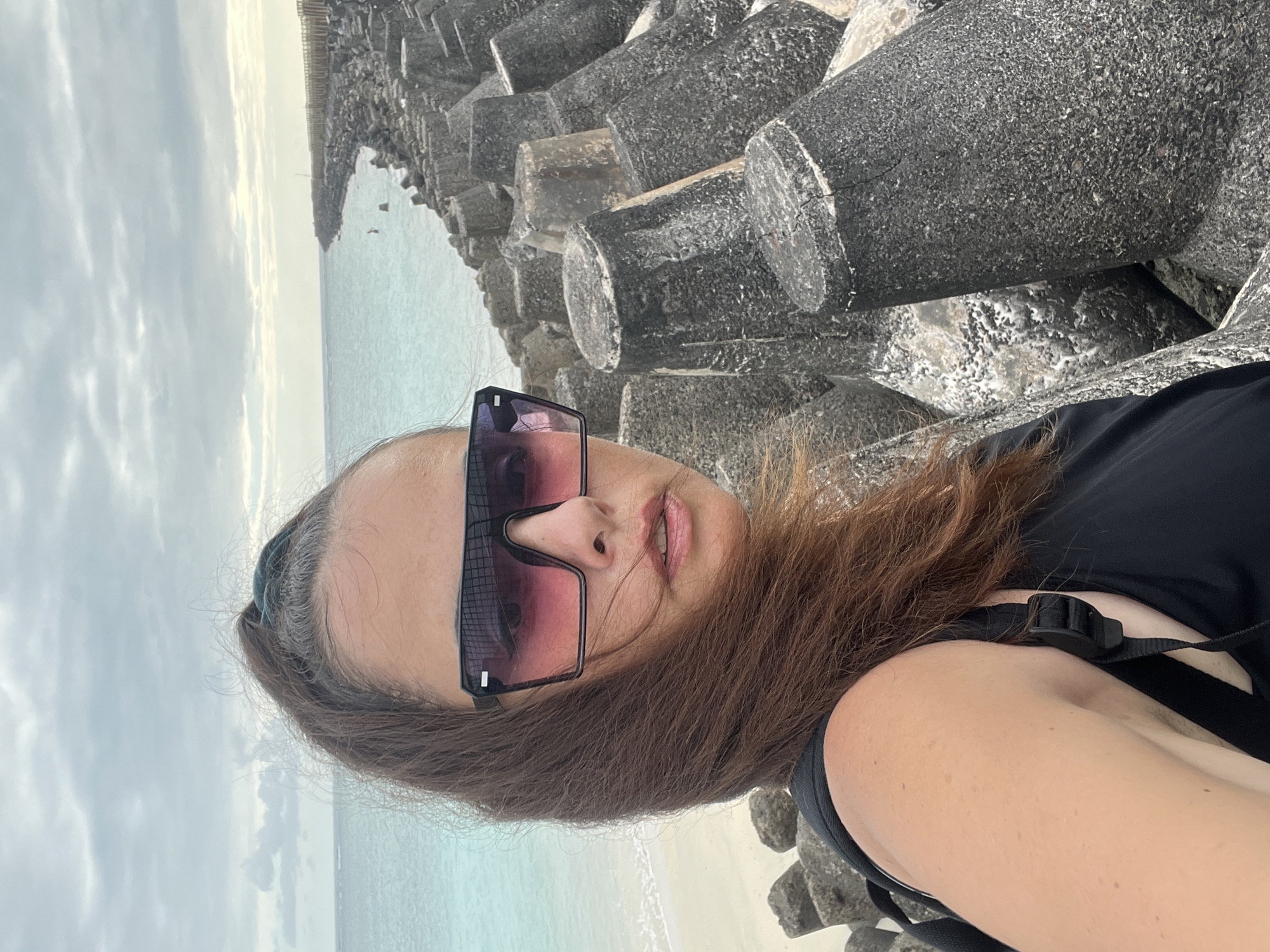 A woman takes a selfie on a rocky shore with a choppy ocean and cloudy sky in the background. The woman is wearing a black top and large sunglasses with pink lenses. She has her hair pulled back in a ponytail and some strands are blowing in the wind. The shore is lined with large, dark concrete barriers. The water is a light turquoise color and there are small waves breaking on a sandy beach to the left of the image. In the distance, there is a pier or walkway with a wooden railing. There are a few small figures visible on the pier and in the water. The overall scene suggests a windy, overcast day at a coastal location.