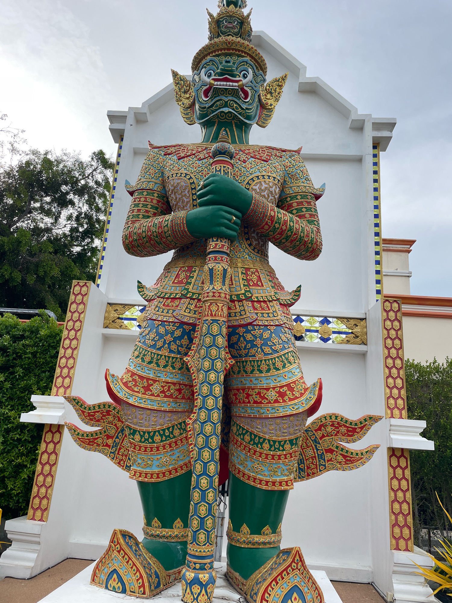 This is a photograph of a large, colorful statue of a green-skinned giant in Pattaya City, Thailand. The statue is elaborately decorated with intricate patterns in red, gold, blue, and other vibrant colors, and it is holding a sword or staff. The giant is positioned in front of a white structure with decorative elements. Lush green foliage and trees are visible in the background, along with a glimpse of another building. The sky is overcast. The statue appears to be a significant landmark or art installation.