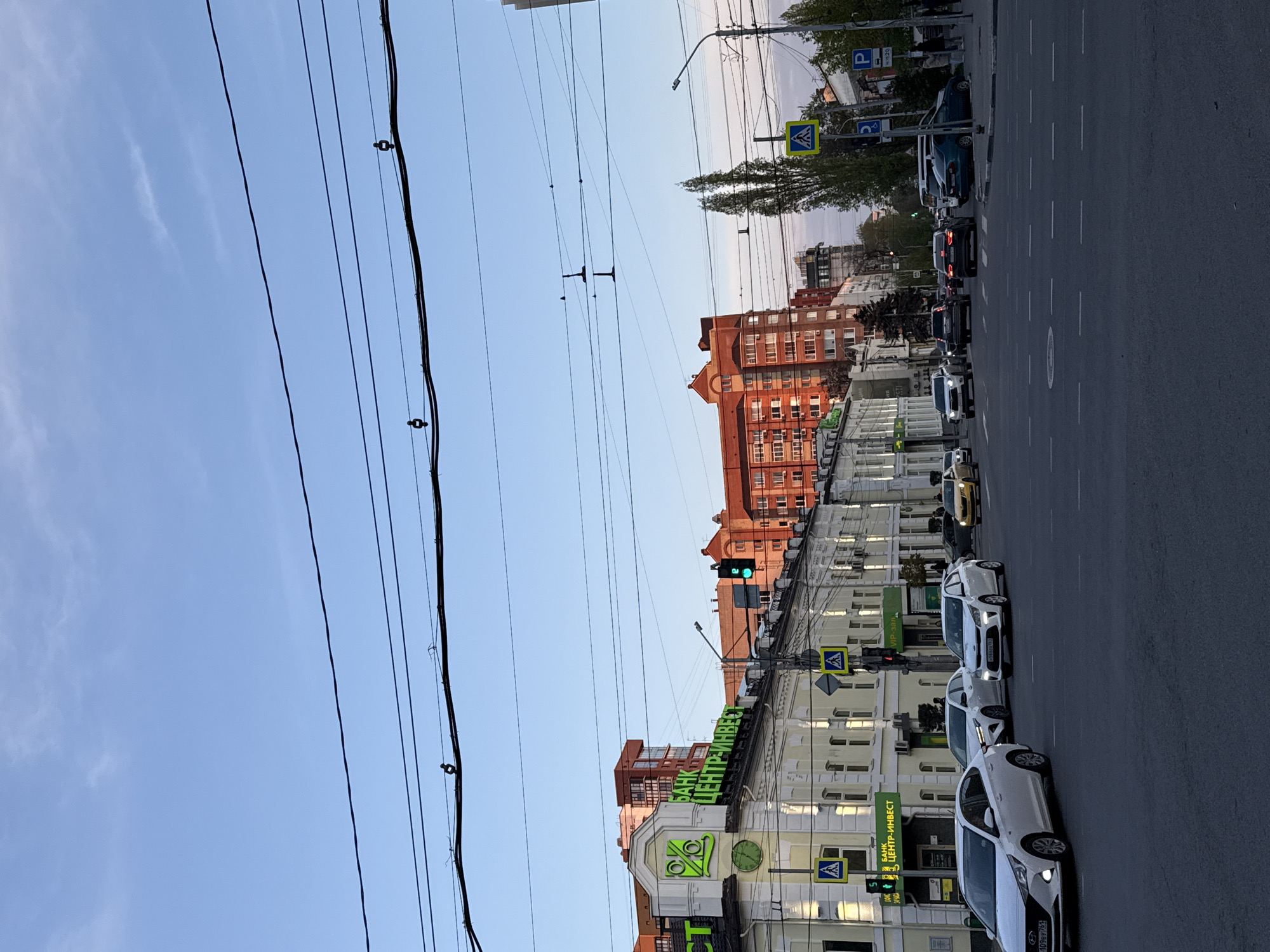 The image depicts a vibrant urban street scene in Rostov-on-Don, Russia, likely captured in the late afternoon or early evening under a clear, bright sky with faint wisps of clouds. The perspective looks down a wide asphalt street, marked with white dashed lines, featuring several cars, mostly white sedans, lined up on the right, suggesting traffic or parked vehicles. A yellow taxi-like car is visible further down the road.
Dominating the upper portion of the frame are numerous black overhead power lines or trolleybus wires crisscrossing against the blue sky, some with small insulators.
On the right side of the street, a row of multi-story buildings lines the road. A prominent light-colored building in the midground features a large green sign with the Cyrillic text "БАНК ЦЕНТР-ИНВЕСТ" (Bank Center-Invest) and a distinctive percentage (%) logo. Below this, a smaller sign reads "VIP-зал" (VIP-hall). Further down the street, a red brick building with many windows adds to the urban landscape.
Visible street furniture includes yellow pedestrian crossing signs (depicting a walking figure on a blue triangle, with the letter 'A'), and parking signs marked with 'P' and '5-20'. A traffic light is visible displaying a green signal, indicating active traffic flow. Sparse trees are scattered along the street and behind the buildings.
The scene captures a typical moment of urban activity, with vehicles present and street infrastructure in use. The Cyrillic text and the style of the license plate "109ey 761" on one of the cars confirm the Russian setting.
