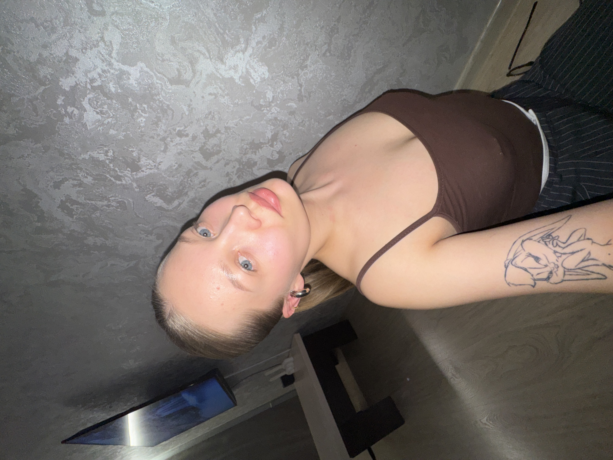A fair-skinned young woman with light-colored hair is lying on her side, partially facing the camera. She is wearing a brown tank top and striped pants. On her left arm is a tattoo. Behind her is a grey wall and the corner of a television. To her right is a lamp. The location is unspecified in the city of Chaikovsky, Russia.