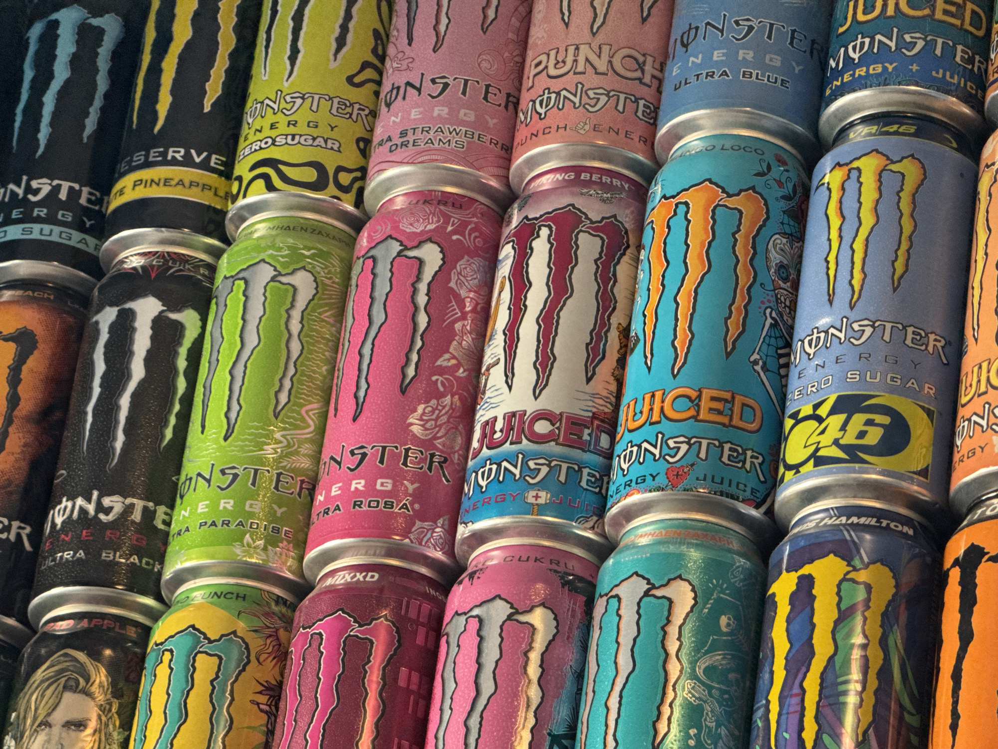 The image displays a close-up, overhead view of a stacked collection of Monster Energy drink cans. The cans are arranged in multiple rows and columns, with some leaning against others. The vibrant colors and distinct Monster Energy logos are prominent on each can. The specific location in the city of Burgas, Bulgaria is not determinable from the image. The scene appears to be a still life arrangement of the beverages.