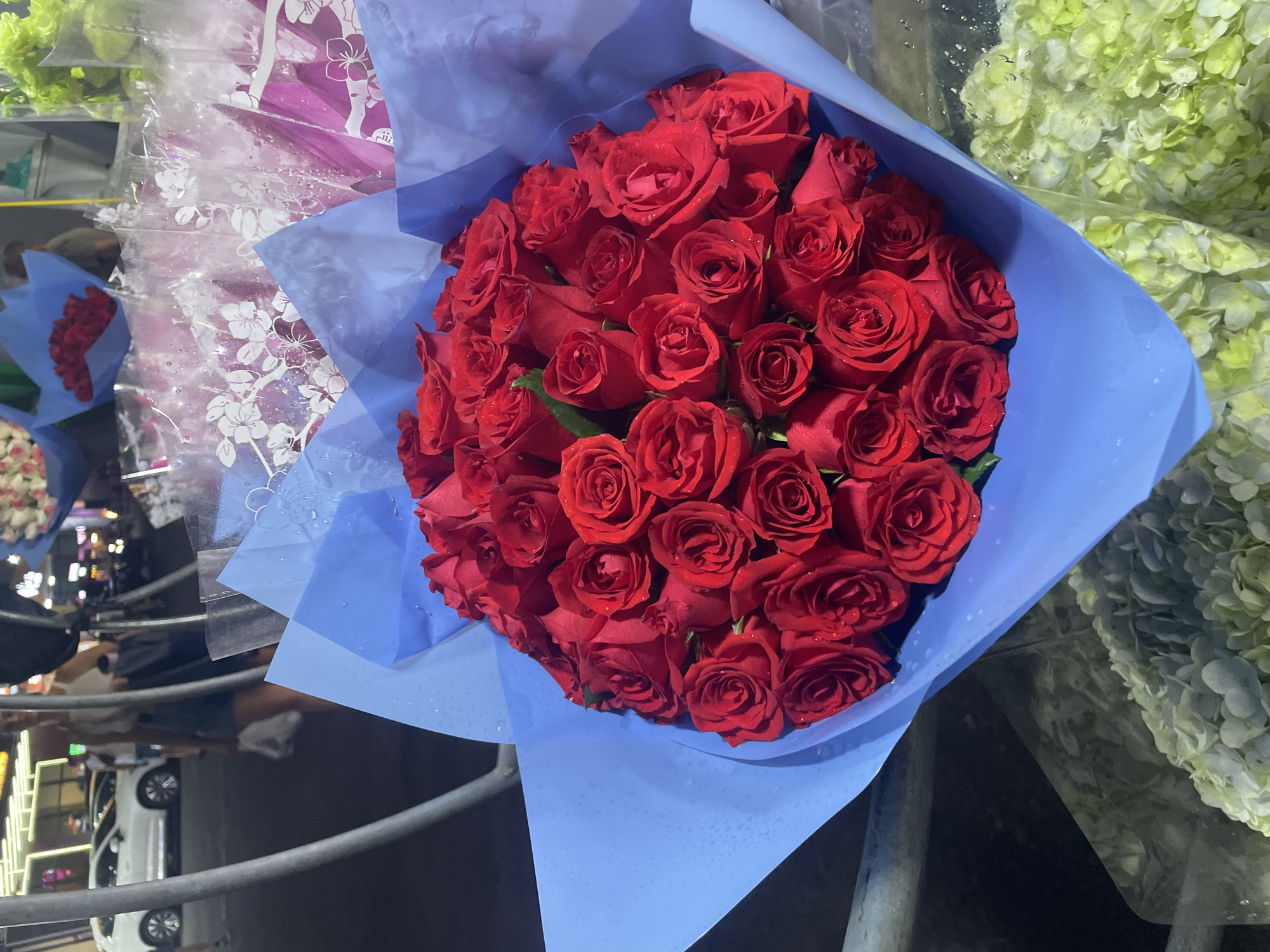 This image captures a vibrant bouquet of red roses wrapped in light blue paper, likely at a flower market or stall in Bắc Nha Trang, Vietnam, at night. The large bouquet is the central subject, prominently displayed and taking up a significant portion of the frame. Numerous red roses are tightly arranged, with visible water droplets on their petals and the light blue wrapping, suggesting either recent watering, dew, or rain.
In the background, other floral arrangements are partially visible. To the right, there's a large mass of light green or white flowers, possibly hydrangeas, also wrapped. To the left, behind the main bouquet, another arrangement with pink and purple hues and patterned clear plastic wrapping can be seen.
The setting appears to be an outdoor or semi-outdoor commercial area. Artificial lighting illuminates the flowers, contrasting with the dark background, which indicates it is nighttime. Blurry figures of people, possibly customers or vendors, are vaguely discernible in the mid-ground on the left. A white car and metal railings are visible in the lower left and bottom parts of the image, suggesting a street or roadside location. No visible text is present in the image.