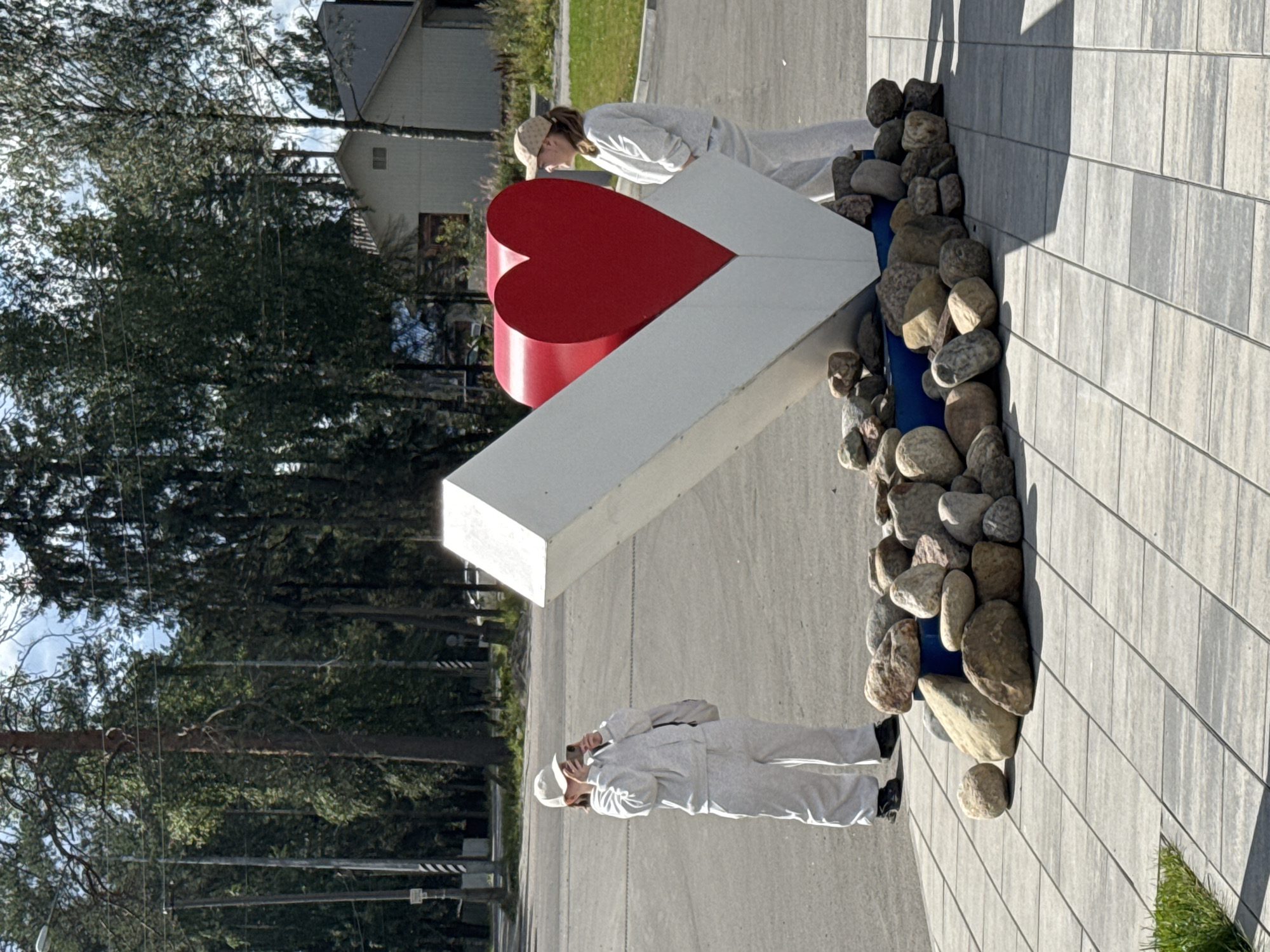 Two individuals, dressed in light-colored tracksuits and baseball caps, are present in the foreground of the image. The individual on the left is holding a mobile phone and appears to be taking a photograph of a large, white, abstract sculpture with a red heart embedded within it. This sculpture is surrounded by a cluster of natural stones. The individual on the right stands near the sculpture, facing away from the camera. The scene is set outdoors, with a paved area in the foreground and a natural environment with trees and buildings in the background. The sky is visible and appears partly cloudy. This monument is located in the city of Lakhdenpokhya, Russia.