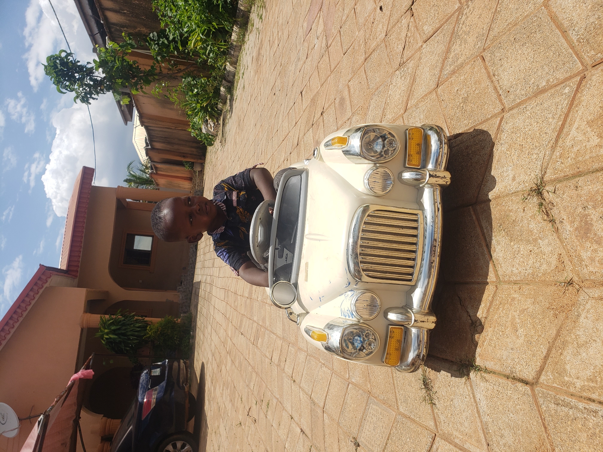 A young Black child sits in a white toy car on a paved courtyard in Sabon Tasha, Nigeria. The child, wearing a dark shirt with a pattern, is facing forward, looking slightly to the left. The toy car is a vintage-style replica, with chrome accents and a prominent grille. In the background, a light brown house with a red-tiled roof is visible, along with a dark blue car parked to the left. The courtyard is paved with light brown hexagonal tiles, and sparse green grass grows in the cracks. A wooden fence and lush green foliage border the right side of the image. The sky is blue with scattered white clouds.