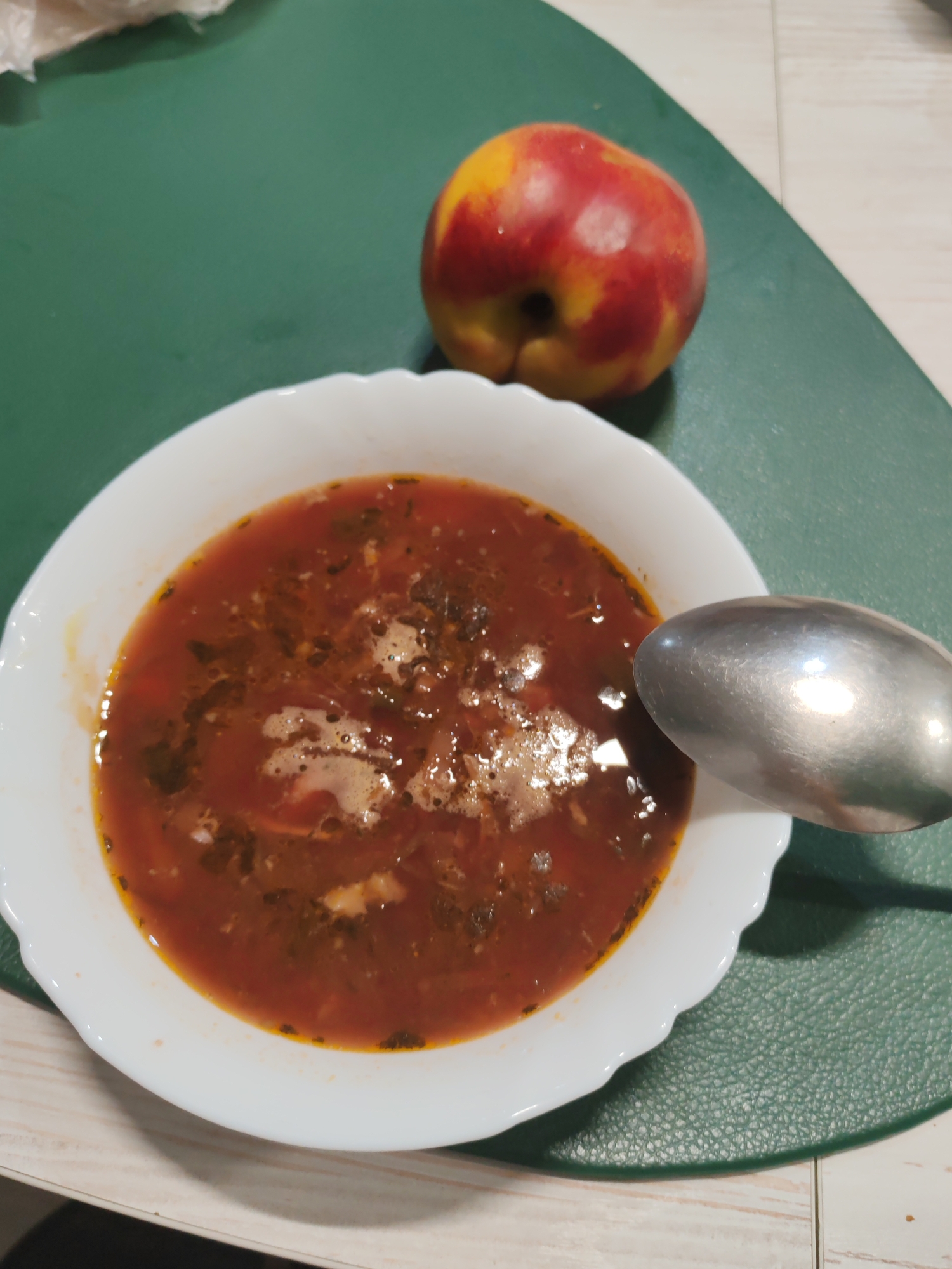 A close-up, high-angle shot shows a white bowl filled with red soup, likely borscht given the visible vegetables and flecks of herbs. A silver spoon rests to the right of the bowl, partially submerged in the soup. Behind the bowl, a nectarine or peach with red and yellow skin sits on a green placemat. The background consists of a textured countertop. The scene appears to be set in a kitchen in Komsomolsk-on-Amur, Russia. There are no people visible in the image.