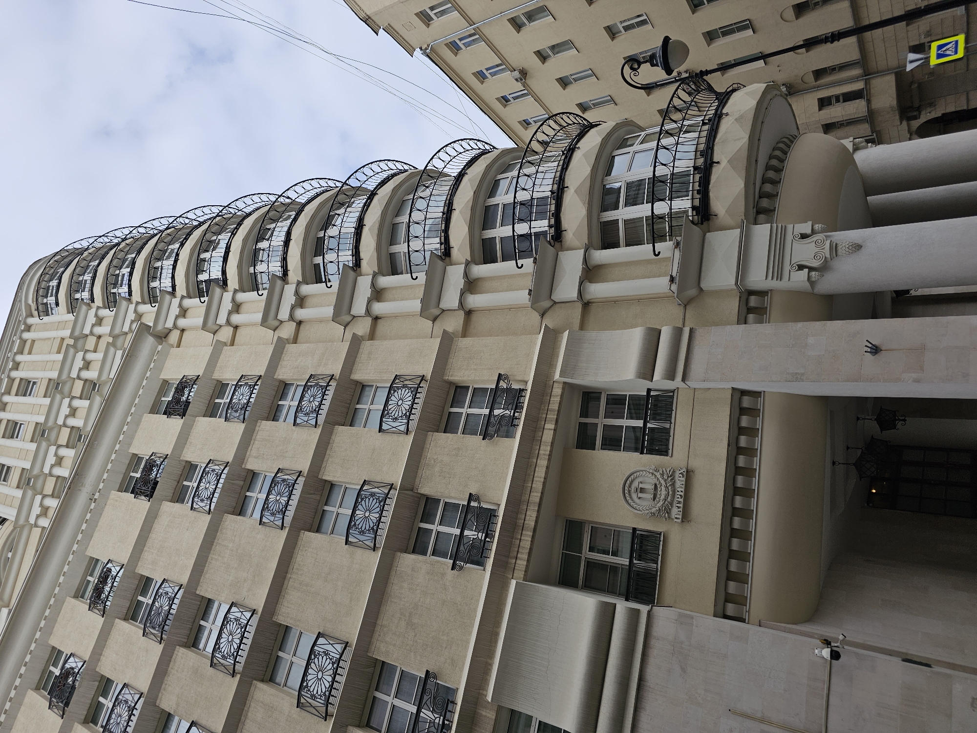 The image shows a close-up, low-angle shot of a building in Moscow, Russia. The building is tall and appears to be residential or hotel, with a light beige facade. It features multiple floors with regularly spaced windows, many of which have decorative black metal balconies with a floral motif. The building has a curved section on the right side, also adorned with balconies. There is an adjacent building to the right, also light beige with windows. A lamppost with an ornate design is visible in the foreground on the right. The sky is overcast with some clouds. The ground level of the main building features an arched entranceway with large columns. There is a security camera mounted on the left side of the entrance. A circular emblem with what appears to be text underneath is visible on the facade above the entrance.