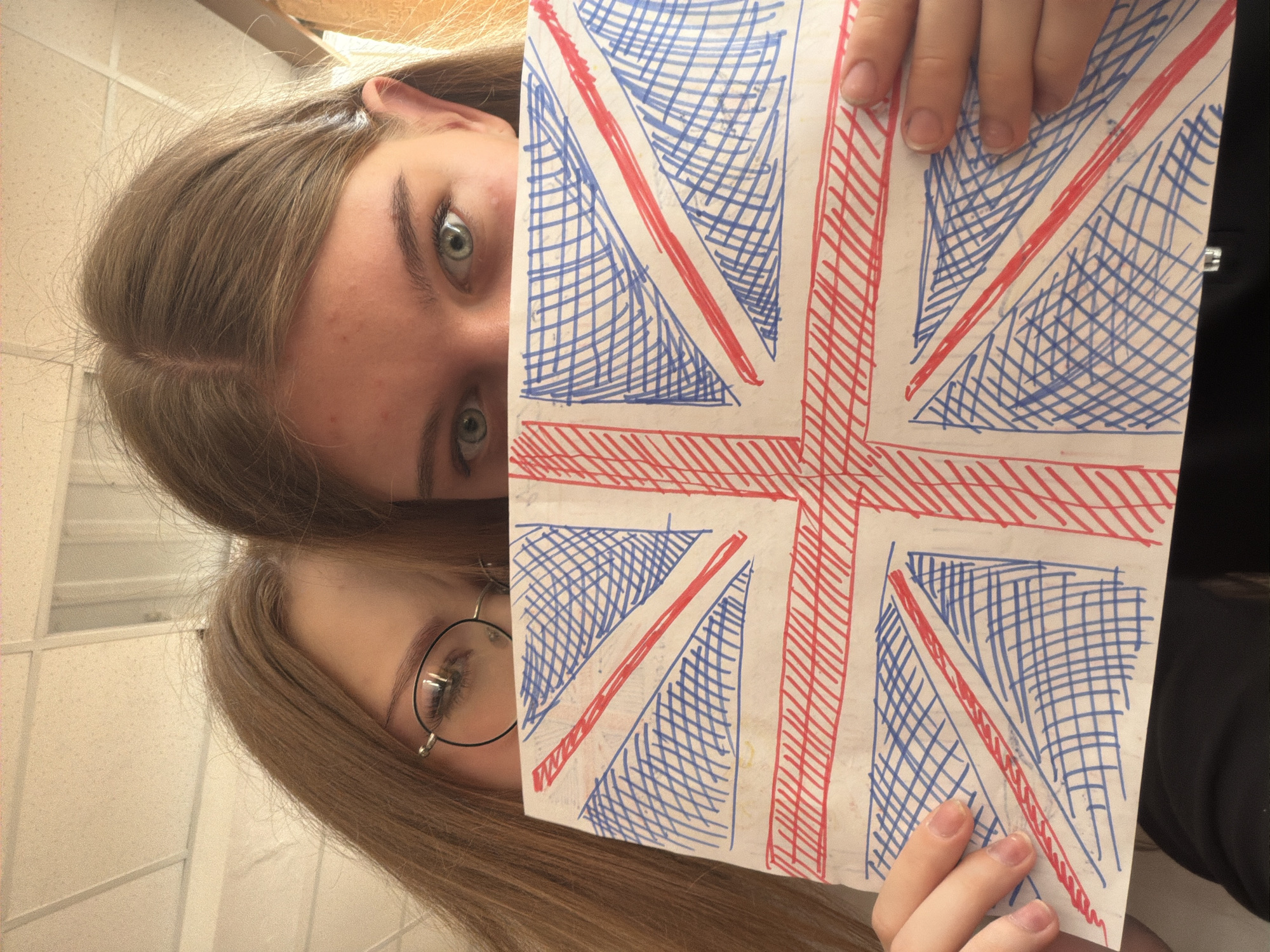Two individuals are positioned at an angle in the frame, holding up a hand-drawn rendition of the British flag. The subject at the top of the image possesses light brown hair, fair skin, and light-colored eyes. The subject at the bottom of the frame is also fair-skinned, with light brown hair and eyeglasses. The setting is an indoor location with a tiled wall in the background. The drawing of the flag is rendered in blue and red ink on white paper, with visible cross-hatching to fill the design. The location is unknown.