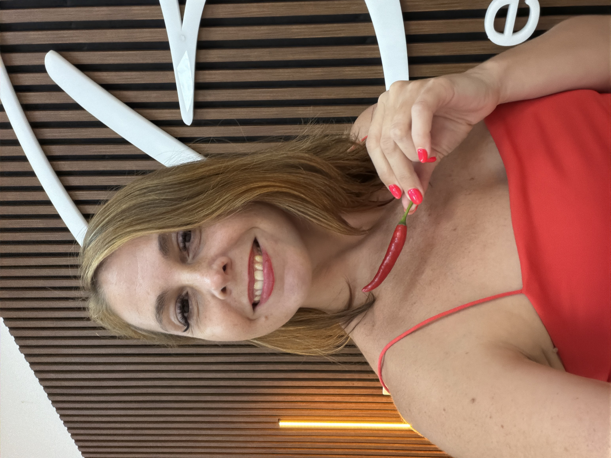 A woman with blonde hair and bright red lipstick smiles at the camera while holding a red chili pepper to her neck. She is wearing a red dress with spaghetti straps. The background is a wall with vertical wooden slats and abstract white shapes. A vertical yellow light fixture is visible on the left side of the frame. The image was taken in Ko Samui District, Thailand.