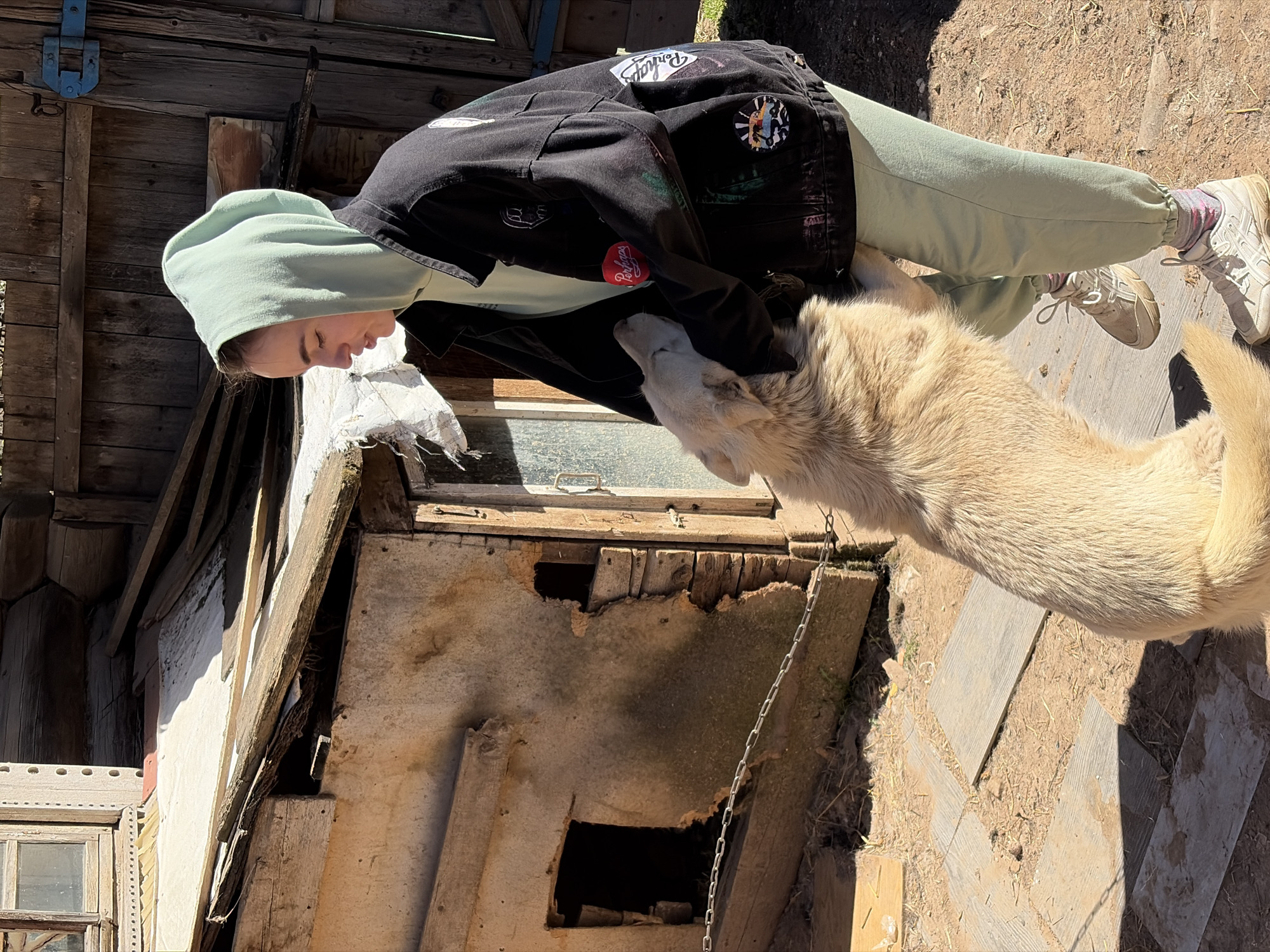 A person with light skin, wearing a light green hoodie, a black jacket with patches, and light green sweatpants with white sneakers is interacting with a large, cream-colored dog near a dilapidated wooden structure. The structure appears to be a shed or small building with missing sections and a weathered window. A chain hangs from the structure near the dog. The ground is a mixture of bare dirt and wood scraps, suggesting an outdoor environment. Based on the geographical metadata, the location is Nizhnii Vishur, Russia.