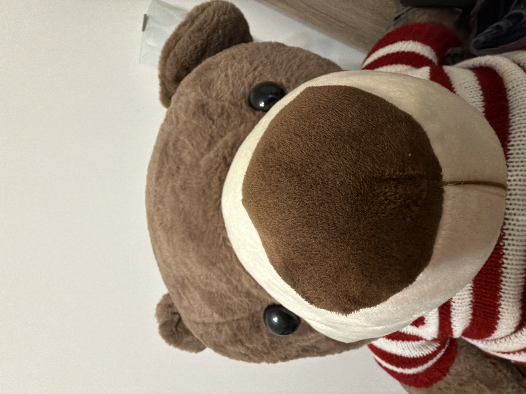 The image shows a close-up of a stuffed animal, specifically a bear. The bear has brown fur, black button eyes, and a large, rounded muzzle that is brown and white. The bear is wearing a red and white striped sweater or scarf. The background is primarily a plain, light-colored surface, likely a wall. The lighting appears even, suggesting an indoor environment. There is no information about location.
