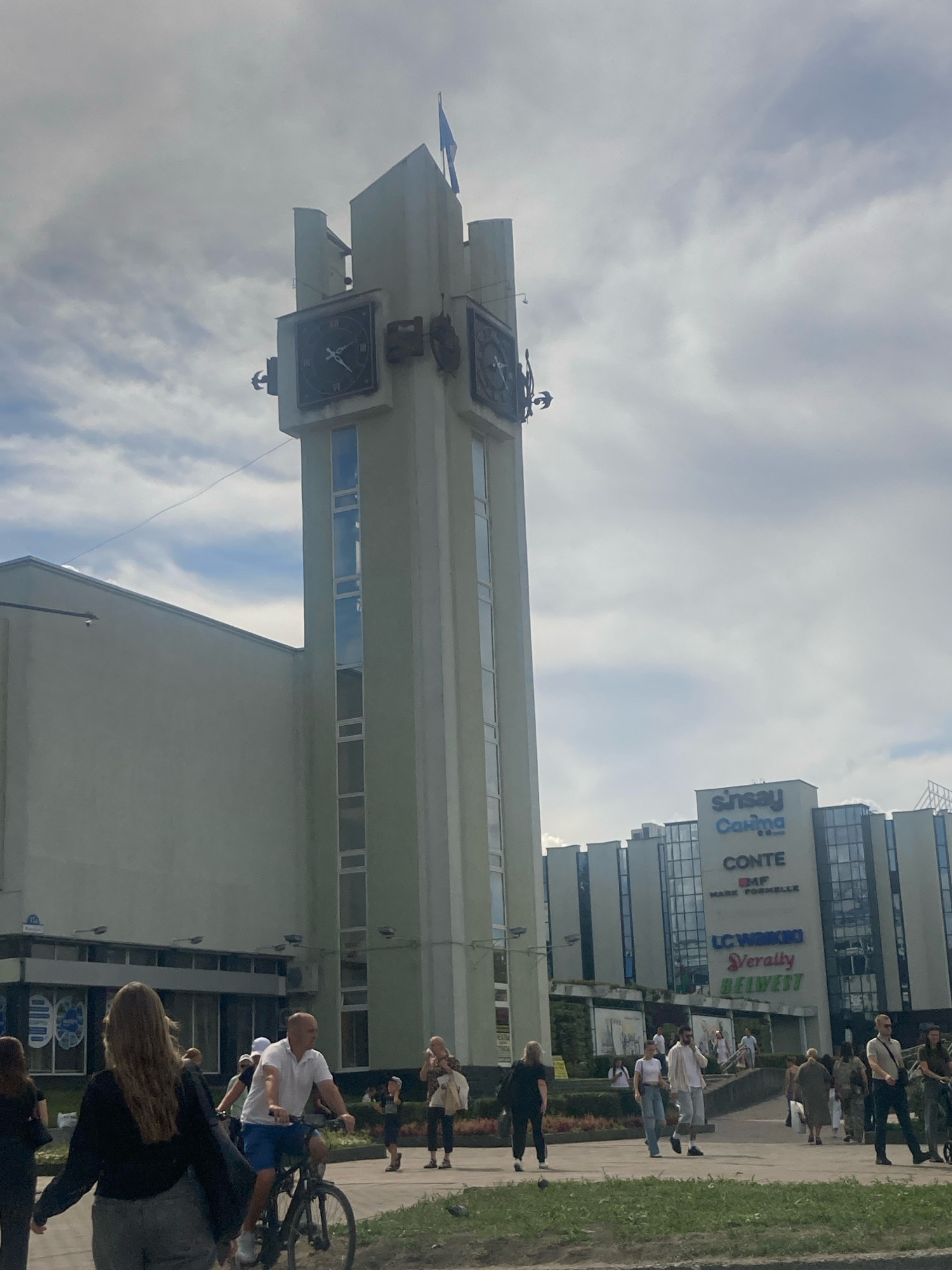 The image depicts a street scene in front of a large building complex in Brest, Belarus. A prominent feature is a tall, light-colored clock tower with two visible clock faces, one showing approximately 1:50 and the other around 1:55. The tower is adorned with decorative elements, including what appears to be a bird sculpture on its left side. To the right of the clock tower, a modern multi-story shopping center is visible, featuring large glass windows and signage that includes "SINSAY," "COSMOSA," "CONTE," "MARK FORMELL," "LC WAIKIKI," "VERALTY," and "BELWEST."
Several people are present in the foreground and mid-ground. A man on a bicycle, wearing a white polo shirt and blue shorts, is riding towards the right. A woman with long blonde hair, wearing a black top and grey pants, is walking away from the camera towards the left. Other individuals are walking, standing, and interacting in smaller groups. The environment appears to be an urban setting with a cloudy sky. The overall impression is of a public space with commercial activity.