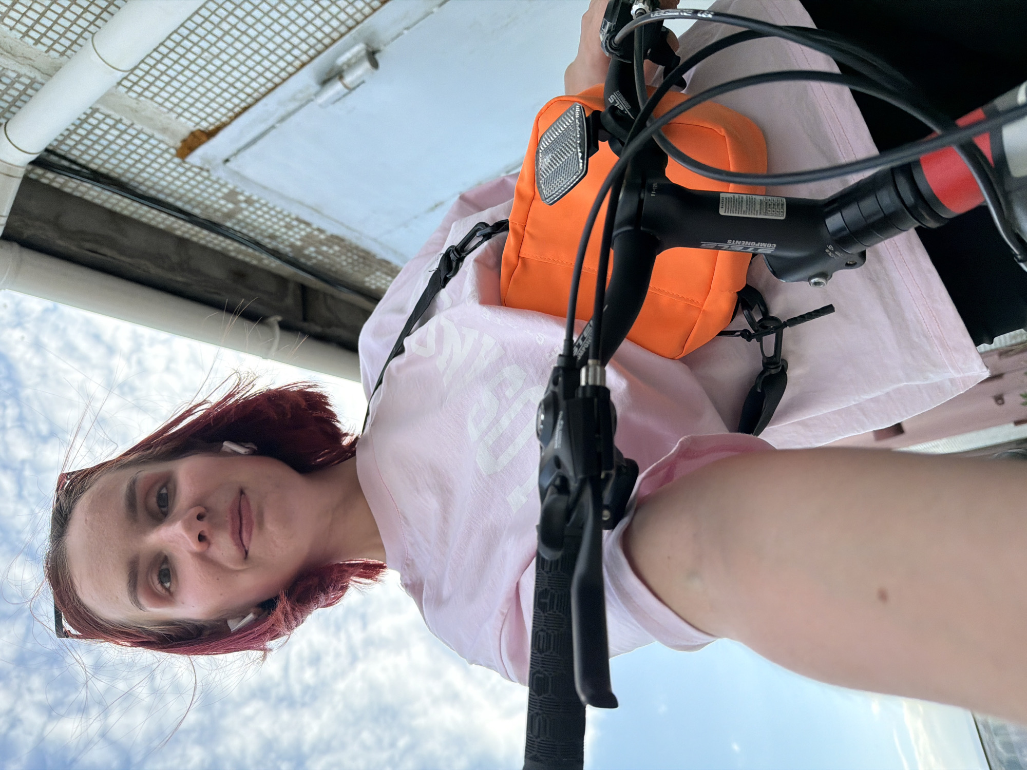 A low-angle selfie shows a young woman with shoulder-length, red hair and fair skin. She wears white wireless earbuds and a pair of sunglasses perched on her forehead. Her facial expression is neutral, and she is looking directly at the camera. The woman is wearing a light pink t-shirt and is holding a bicycle. An orange bag is attached to the handlebars of the bike. The background is split between the sky with scattered clouds and a metal structure overhead, suggesting the photo was taken on a bridge or raised platform. While specific details about the location in Chaikovsky, Russia cannot be confirmed with the provided information, it appears to be an outdoor public space.
