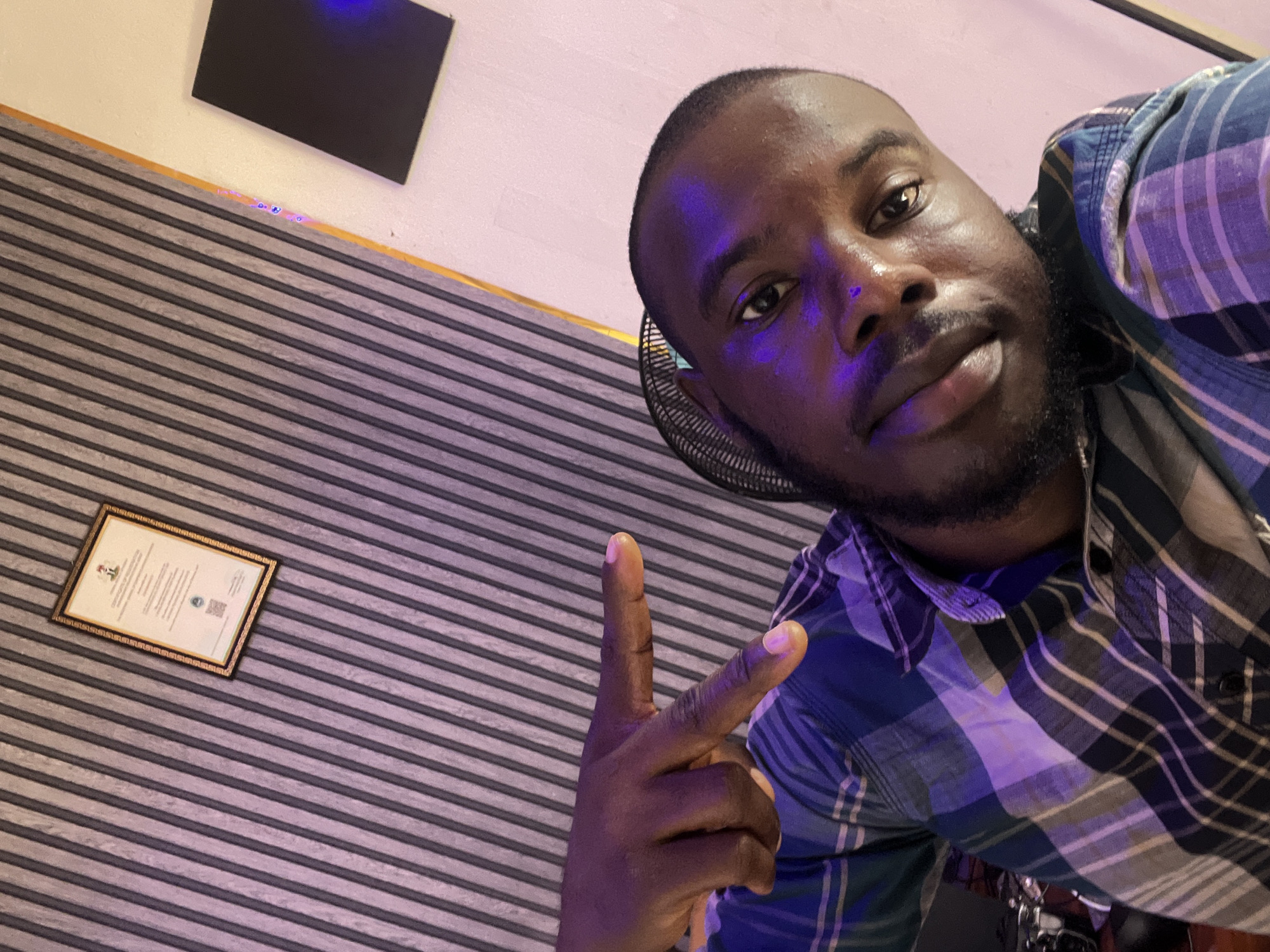 The image is a selfie taken by a man in what appears to be a studio or a room with soundproofing panels on the wall. The man is wearing a blue and plaid shirt. He is pointing with his right hand towards the camera. Behind him, there is a framed certificate on the wall and a black panel. The lighting in the room is a mix of purple and pink hues, possibly from studio lights. The overall impression is of a casual, personal snapshot. There is no information in the image to indicate a specific location in the city of Jos, Nigeria.