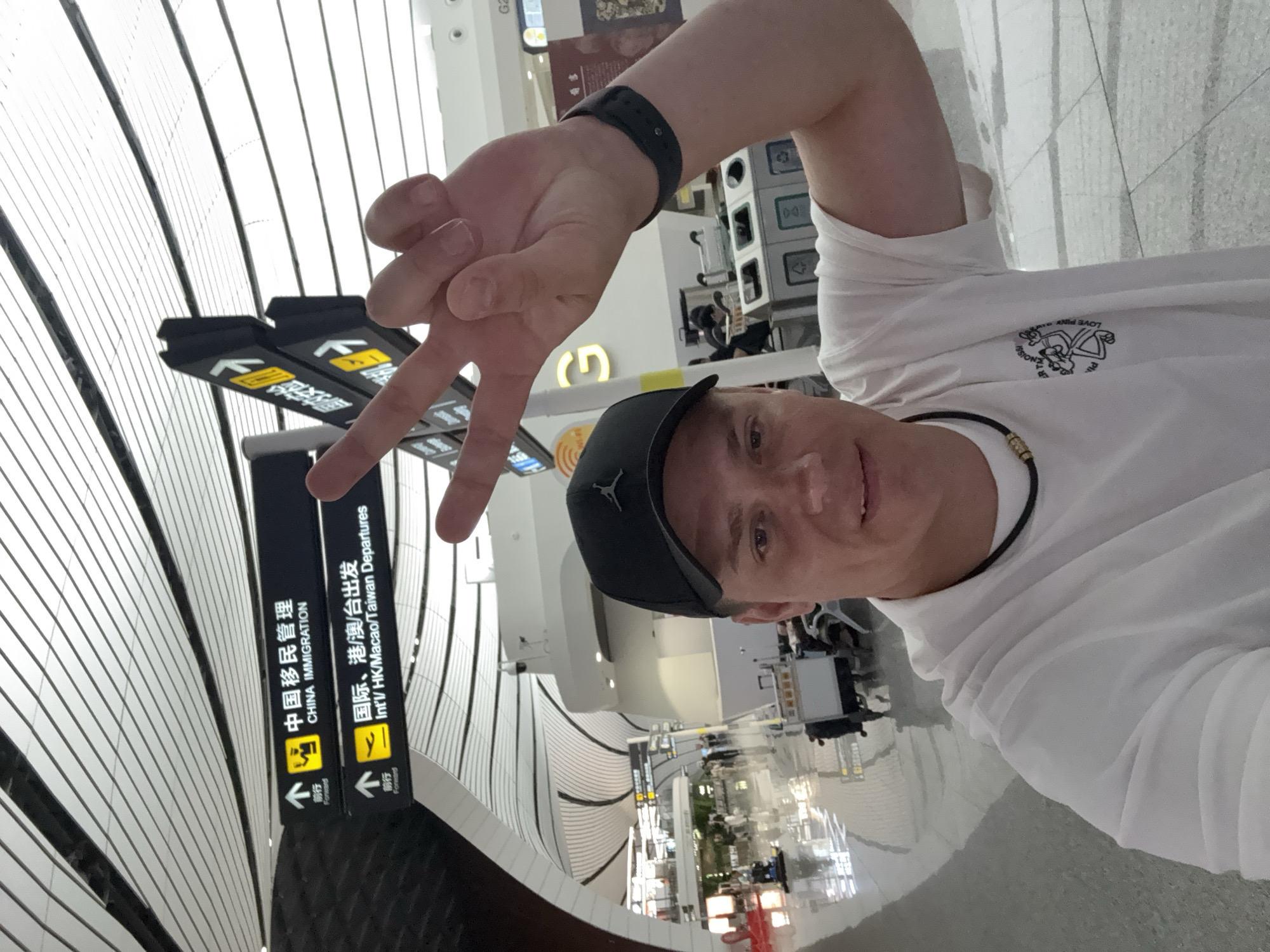 A man in a white t-shirt and a black baseball cap is taking a selfie in an airport terminal. He is holding up two fingers in a peace sign. Behind him, signs indicate "CHINA IMMIGRATION" and "Int'l HK/Macao/Taiwan Departures". The airport appears modern with a distinctive ceiling design. The man is in the city of Guangyang District, China.