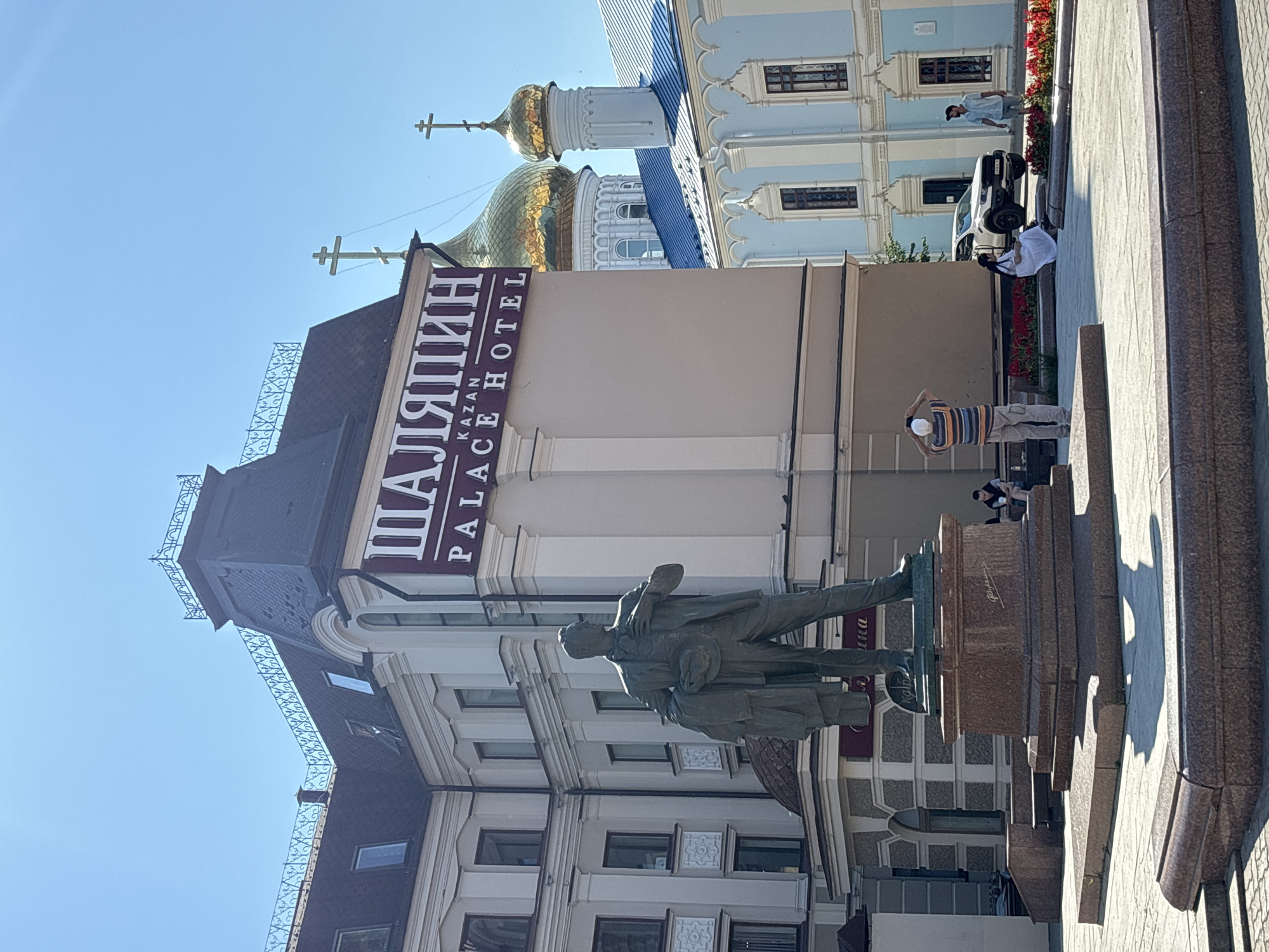 This is a medium-shot, eye-level, outdoor photograph taken on a sunny day in Kazan, Russia, likely in the vicinity of the Kazan Palace Hotel. In the foreground, a bronze statue of a man in a suit holding a coat and hat stands on a multi-tiered granite pedestal. The statue is positioned in a paved square. Behind the statue, the Kazan Palace Hotel building is visible, with a prominent sign in Russian and English that reads "ШАЛЯПИН KAZAN PALACE HOTEL". To the right of the hotel, a light blue building with ornate white trim and a blue roof is visible, likely part of a church or religious complex, with golden domes and crosses crowning its spires. In the middle ground, several people are present. One individual stands facing the hotel, holding up a white hat or phone, possibly taking a photograph. Another person is seated on a bench with red flowers in front of them. Further back, a white SUV is parked, and two individuals are walking. The sky is clear and blue.