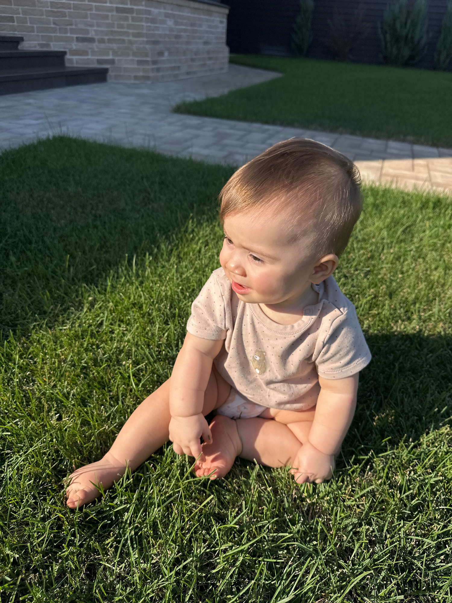 A Caucasian baby is sitting on a bright green lawn. The baby is wearing a light brown onesie with a small bear graphic and possibly a diaper. The baby is facing to the left and looking slightly downwards. The baby's right leg is bent with the foot resting on the ground, and the left leg is crossed over the right. The baby is touching the grass with their right hand. The background includes a brick wall with stairs, a paved pathway, and more grass with some trees. The sun is casting a shadow on the grass to the right of the baby. The overall environment appears to be a backyard or garden area.