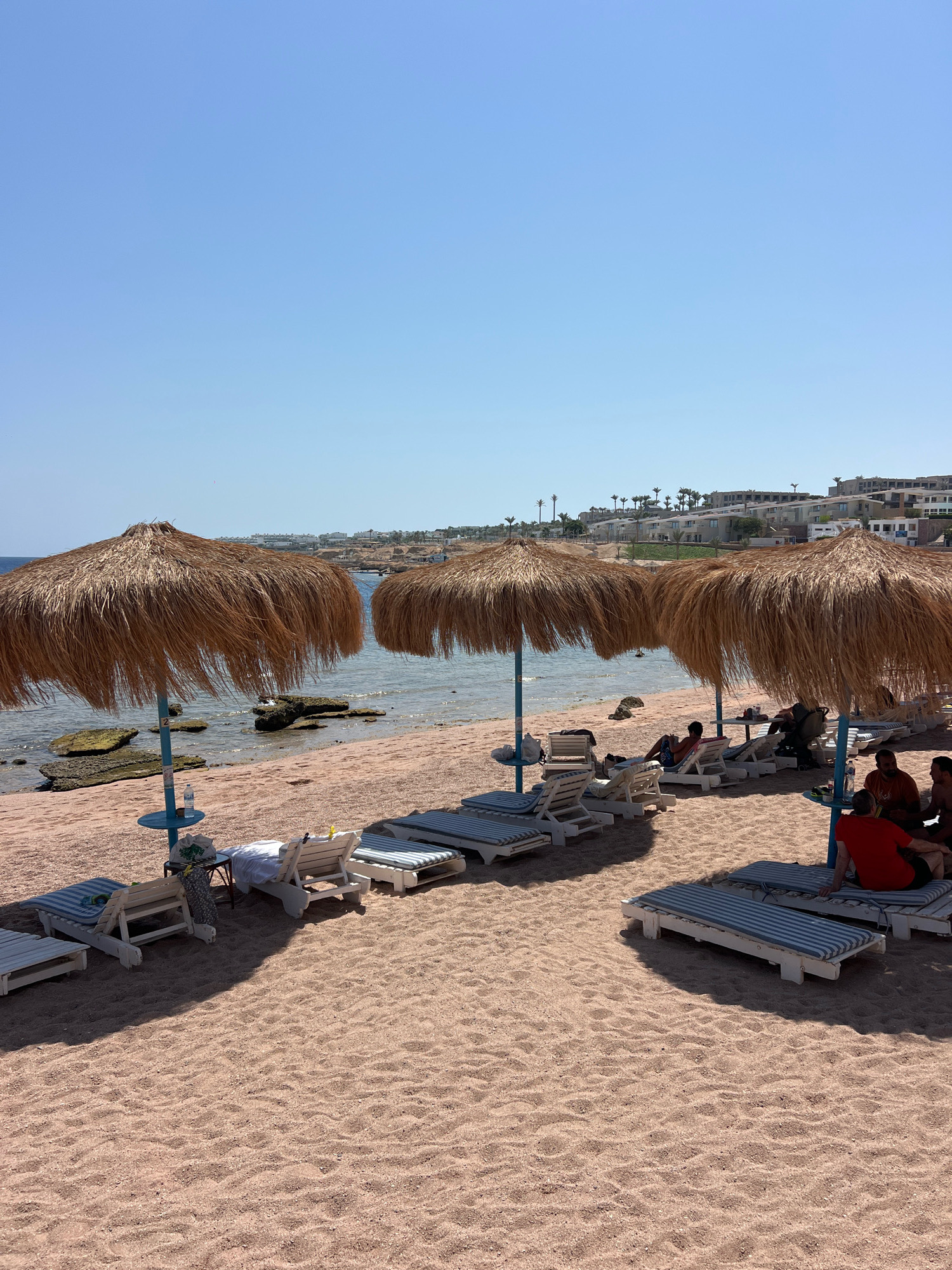 This image captures a serene beach scene in Sharm El Sheikh, Egypt, on a clear, sunny day. The foreground is dominated by a sandy beach, with the fine, light-pink sand showing clear footprints from previous visitors. Several lounge chairs with blue and white striped cushions are arranged on the sand, some empty and others occupied.
Three large, thatched umbrellas provide shade. The umbrellas are made of dried palm fronds and are supported by blue poles. Beneath one umbrella, a small blue table holds a bottle of water and a bag.
The sea is calm and shallow, with clear, sparkling blue water. Several large rocks are visible in the water close to the shore, some covered with green algae. In the distance, a line of buildings, likely hotels or resorts, with white and beige facades and palm trees, stretches along the coastline. The sky above is a vast expanse of clear, pale blue.
Several people are present on the beach, relaxing on the lounge chairs. Some are seated, while others appear to be lying down. One man in a red shirt is seated on a lounge chair, facing away from the camera. Another group of men are gathered near a table, engaged in conversation. The atmosphere is peaceful and suggests a typical tourist experience at a beach resort.