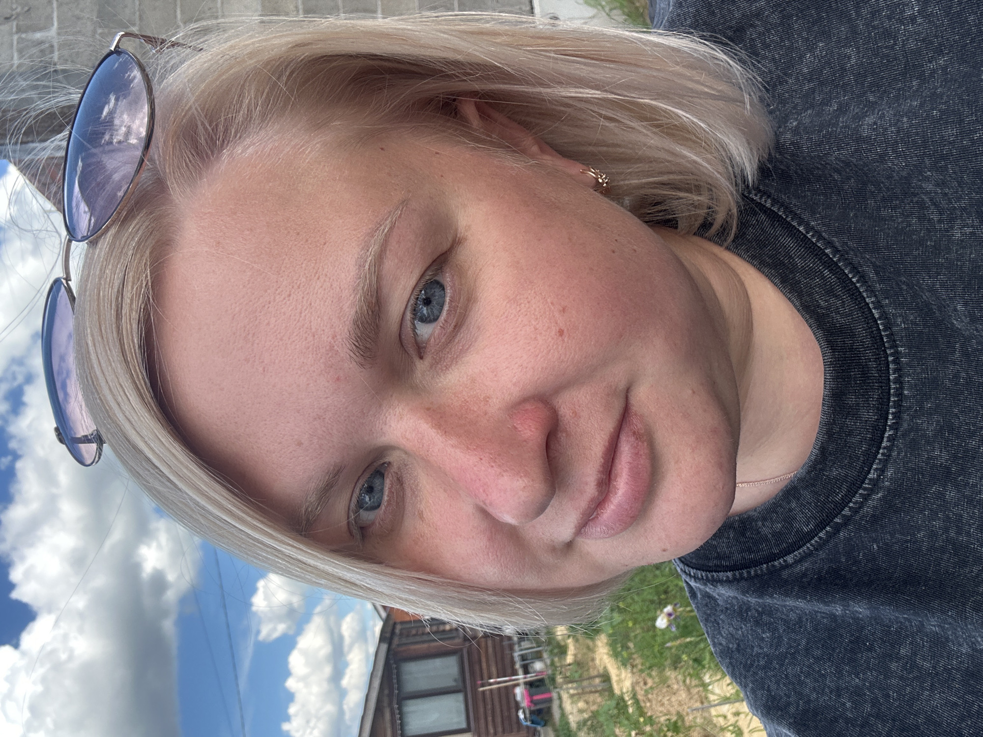 This is a vertical, close-up selfie of a young Caucasian woman with blonde hair, taken outdoors. She is wearing a dark gray t-shirt and sunglasses perched on her head. Her face is turned slightly towards the camera, and her blue eyes are visible. The background shows a bright blue sky with white clouds, and a glimpse of a wooden building and green grass, suggesting a rural or suburban setting. The lighting indicates it is daytime, likely sunny, given the bright sky and reflections in the sunglasses. The woman appears to have a neutral or slightly smiling expression. There is no visible text in the image. The location context provided is Chaikovsky, Russia.