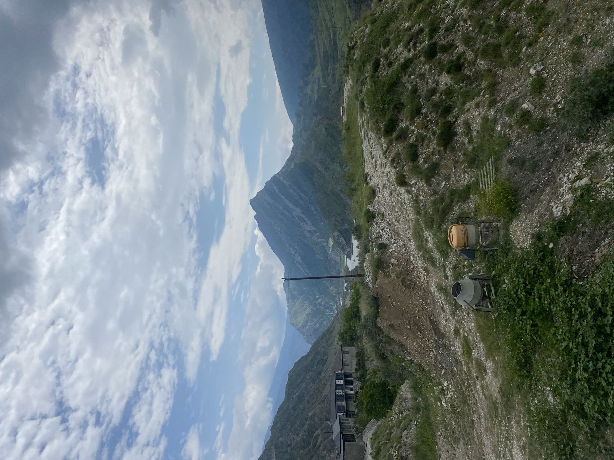 The image shows a mountainous landscape with a cloudy sky. In the foreground, there are two concrete mixers on a grassy, rocky slope. Behind the concrete mixers, there is a dirt path and some vegetation. Further in the background, there are several buildings, a river, and more mountains. The scene appears to be in or near the village of Chalda, Russia, based on the architectural style of the buildings and the mountainous terrain. The lighting suggests it is daytime, with the sun partially obscured by clouds.
