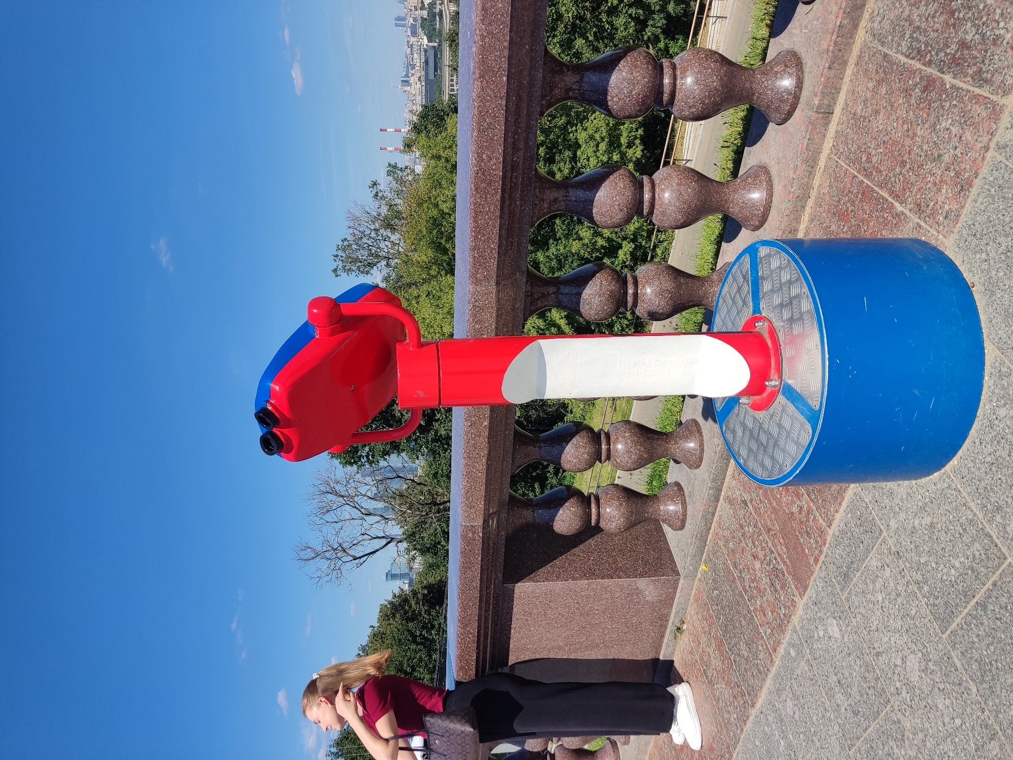 A young woman with blonde hair pulled back in a ponytail, wearing a maroon t-shirt and black pants, stands on a stone terrace overlooking a city skyline. She is holding her hair with her right hand, her eyes closed, and her head tilted back. To her right is a red and blue coin-operated viewing machine. The terrace has a granite railing with decorative balusters. In the background, the cityscape of Moscow, Russia, is visible with trees and buildings under a clear blue sky.