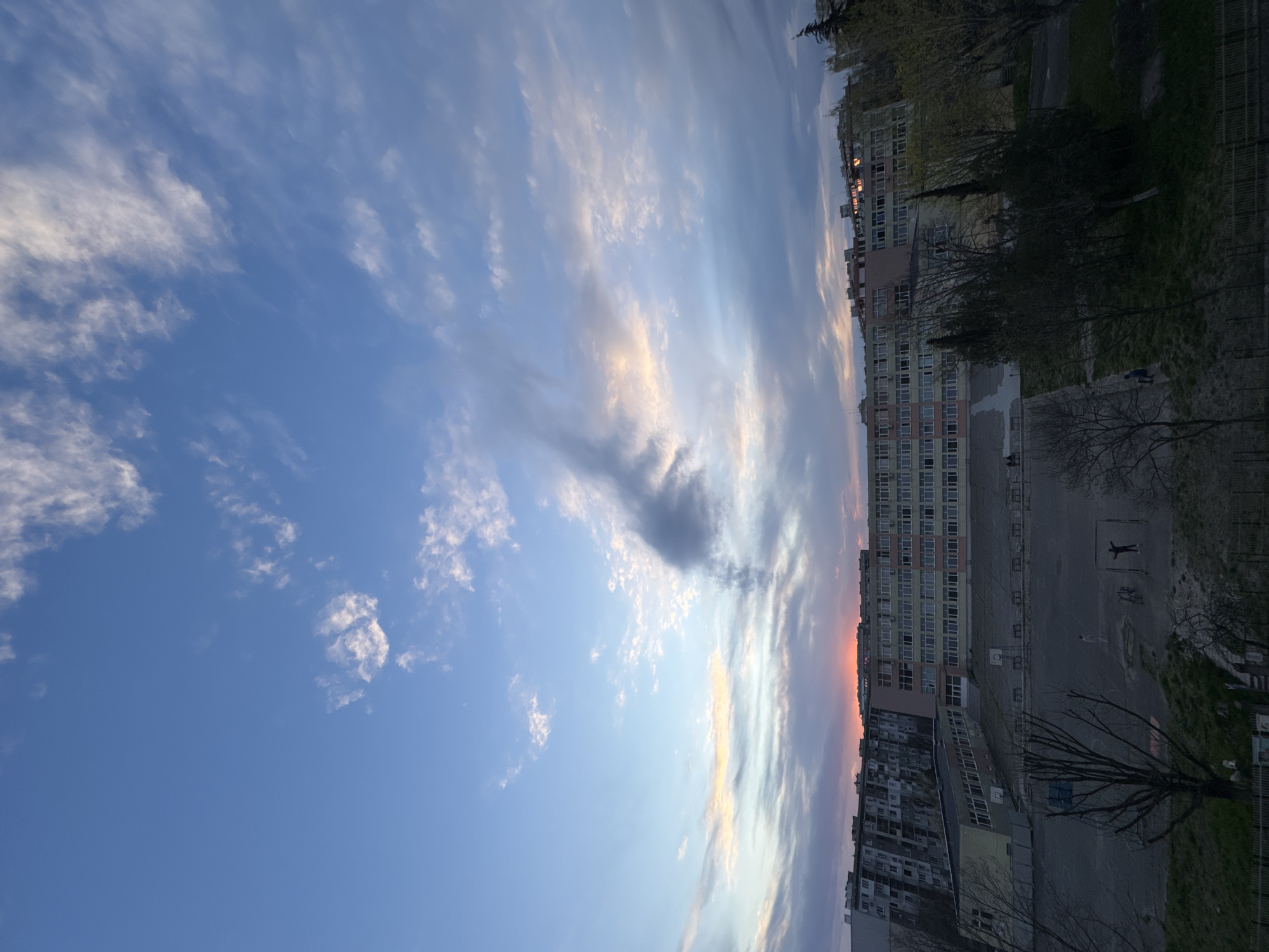 The image captures an outdoor scene in a city, likely in Bulgaria, as indicated by the city's name Burgas.
**Environment:**
The sky dominates a large portion of the image, displaying a gradient of blue and white, with streaks of clouds and sun rays.
There are urban elements, with multi-story buildings visible along the right.
**People:**
There are two people in the playground.
**Objects:**
The image includes buildings, possibly apartment complexes, trees with bare branches, and a basketball playground.
**Actions:**
People are playing in the playground.