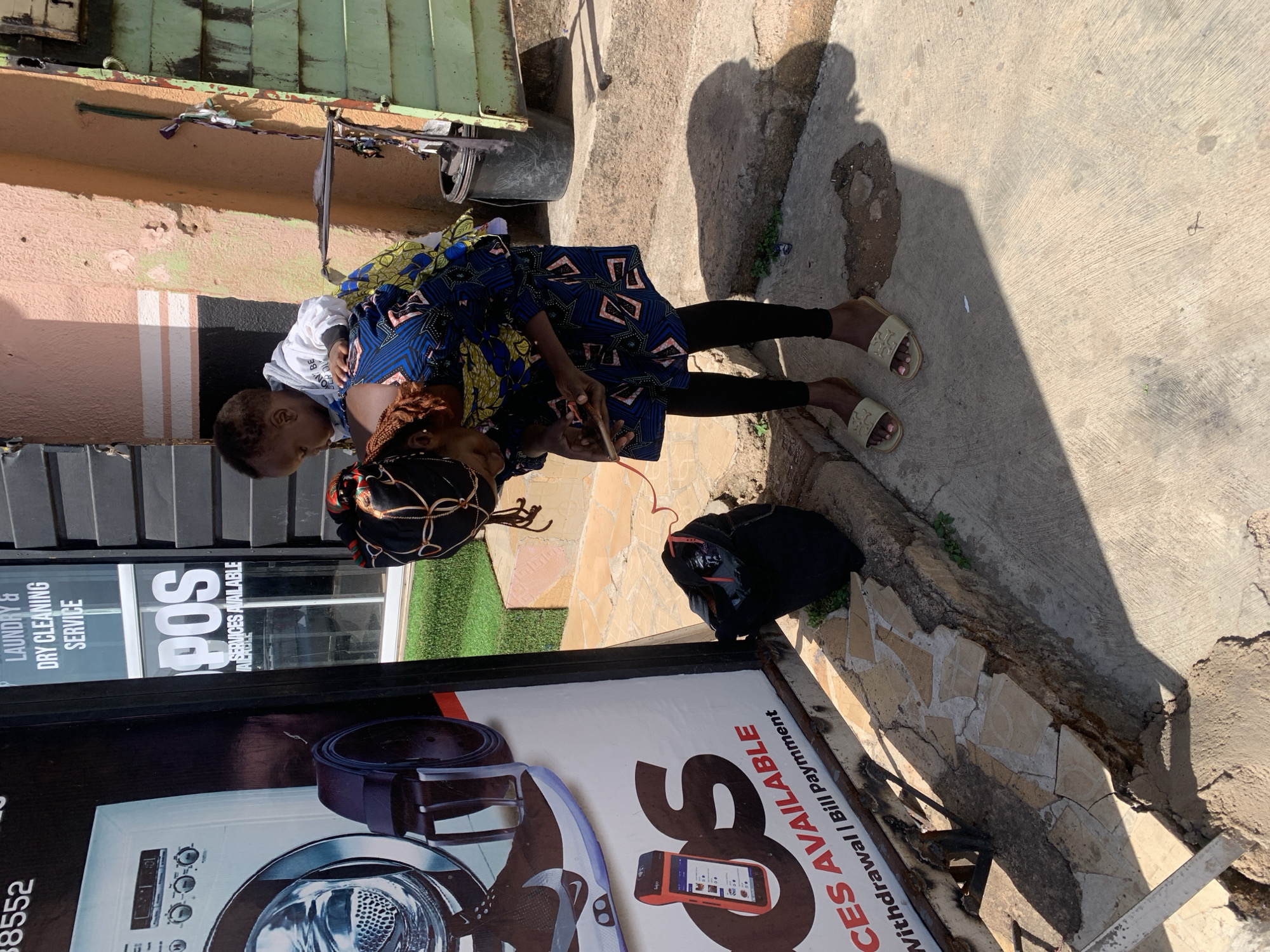 A medium, long-shot photograph depicts a woman holding a child in front of a shop. The woman is wearing a patterned blue dress and black leggings. She is also wearing sandals and a headscarf. The child is wearing a white top. The woman is holding a mobile phone and seems to be doing a transaction. Next to her is a black bag. The shop's sign advertises laundry and dry cleaning services, as well as POS services including withdrawal and bill payment. The shop has a large advertisement featuring a washing machine. The setting appears to be in a city, possibly Jos, Nigeria, given the style of dress of the woman and the types of services advertised on the shop signage. The ground is concrete and there is a step next to the sidewalk. There are buildings in the background. The light suggests the photo was taken during the day.
