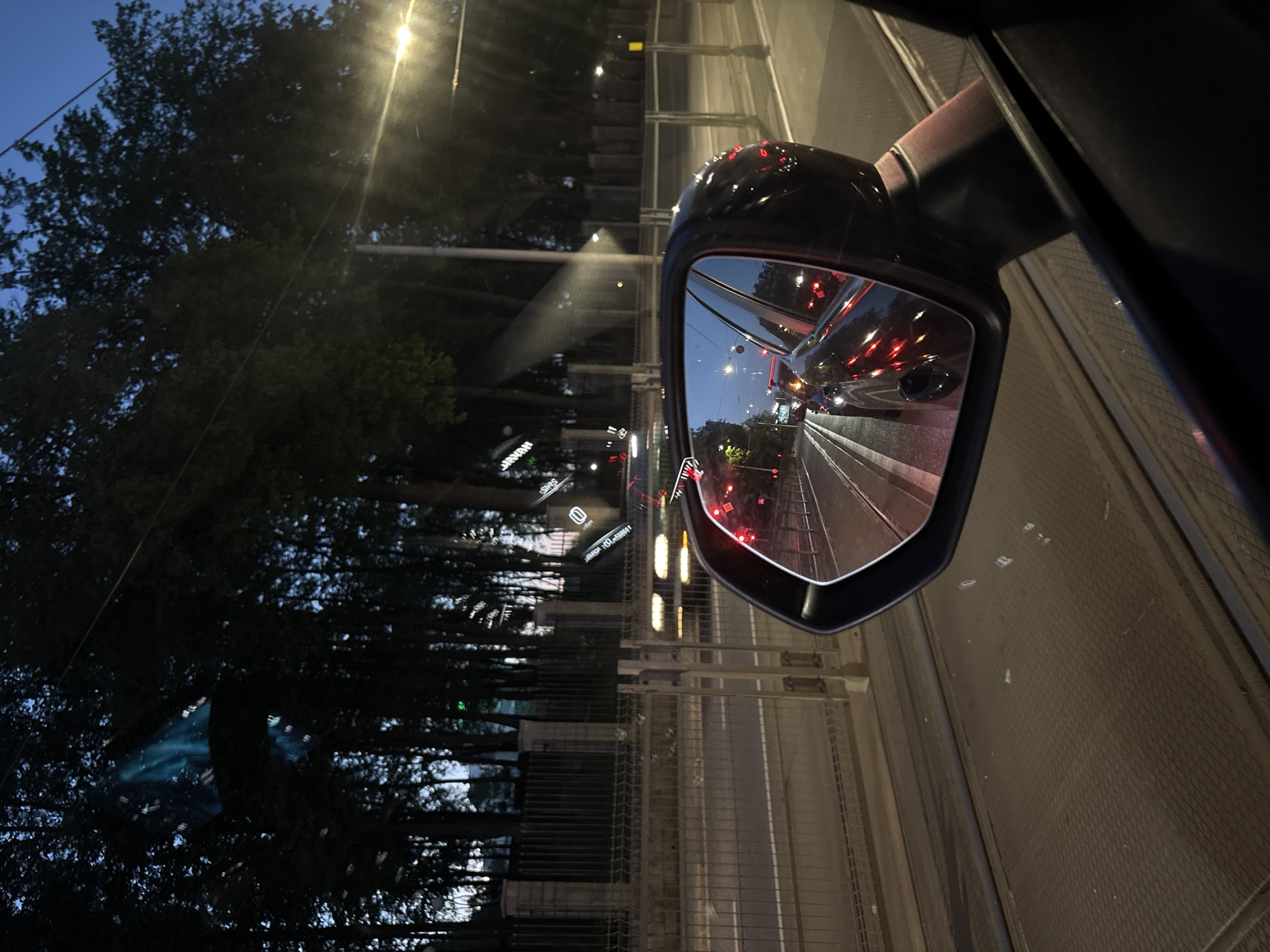 The image shows a view from a car's side mirror at night. The mirror reflects a lane of traffic with red taillights of vehicles. Outside the car, a fence and trees line the road. Streetlights illuminate the scene. Based on identifying the "Swiss Krono" advertising along with the "100-leciu" signs, the location is Minsk, Belarus.