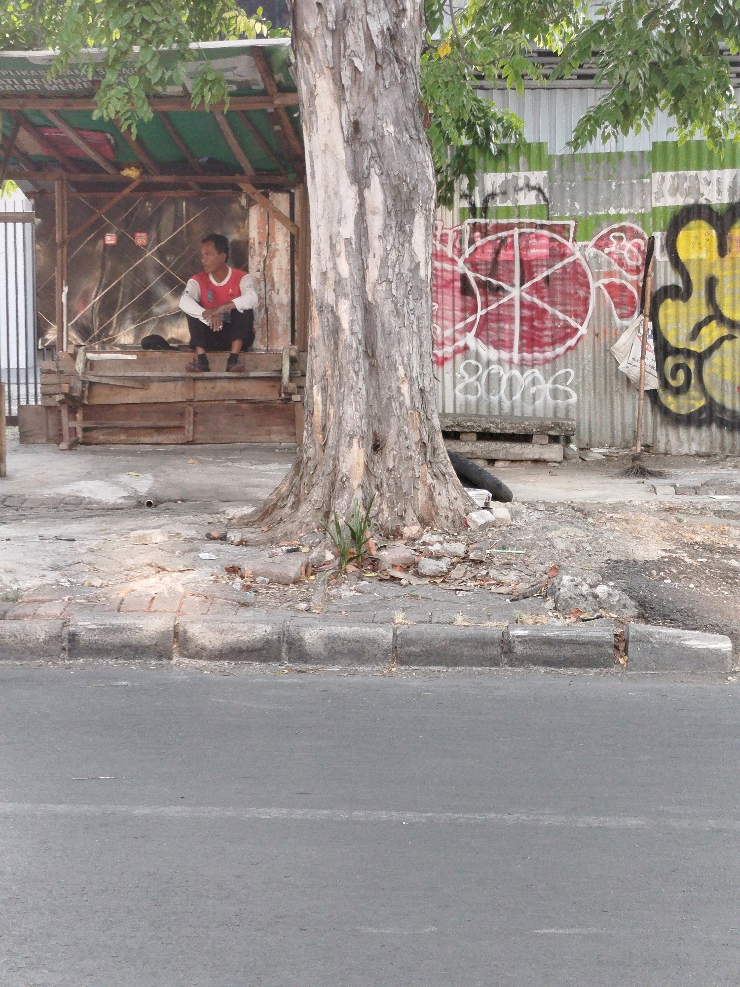 An outdoor, medium shot captures a man sitting on wooden steps in a roadside shelter. He is wearing a red and white long-sleeved shirt and black pants. To his left, on the ground, is a black bag. The shelter has a green roof and is constructed with wood and corrugated metal. Behind the shelter, a corrugated metal fence is covered with graffiti, including red circular designs and yellow abstract shapes. A large tree trunk is positioned in front of the fence and shelter, to the right of the man. The ground in front of the shelter is paved with concrete and bricks, and debris such as leaves and rocks are scattered around. A paved road with a white dividing line is visible in the foreground. The overall impression is of an urban environment with signs of informal habitation and street art. The scene is located in Surabaya, Indonesia, as indicated by the text visible on a sign within the shelter.