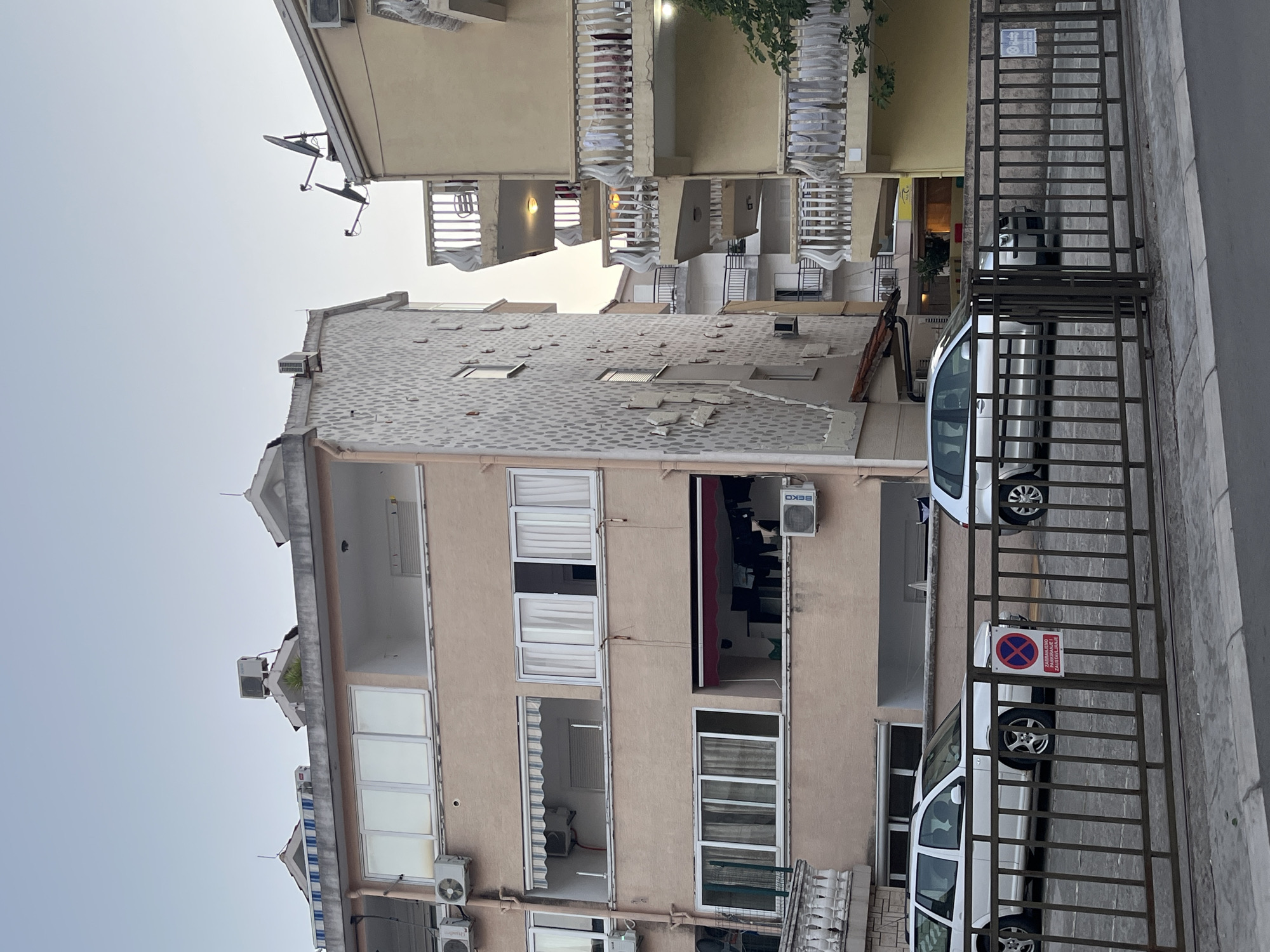 The image shows an exterior view of apartment buildings in Budva, Montenegro, at dusk. In the foreground, a metal fence encloses a parking area where two white cars are parked. One is a station wagon and the other appears to be a hatchback. A circular red and white sign indicating "No Parking" is visible on the fence.
The buildings are multi-story, with balconies on the right-hand building. Air conditioning units are mounted on the exterior walls of the buildings. Satellite dishes are visible on the roof of the yellow building on the right. Clothes are hanging on a clothesline on a balcony of the pink building on the left. The sky is clear and a pale blue, suggesting the sun has recently set. The overall scene is residential and urban.