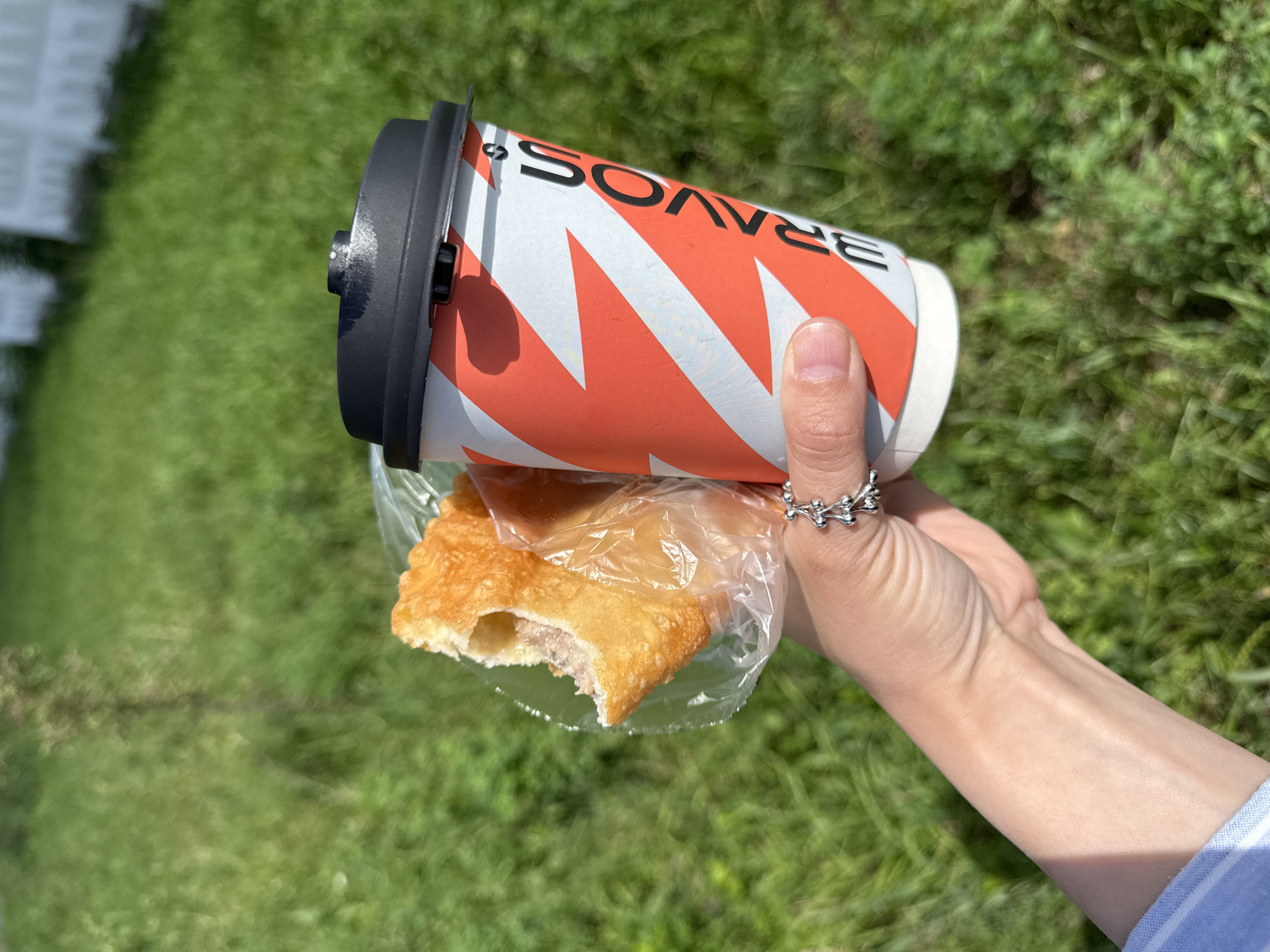 The media file is a close-up shot of a hand holding a coffee cup and a pastry. The coffee cup is white with orange zigzag patterns and black text that reads "BRAVOS". The pastry is golden brown and appears to be a type of fried turnover, with a bite taken out of it, revealing a meat filling. The hand is adorned with a silver ring that has a nature-inspired design.
The background is a blurred green grassy area, suggesting an outdoor setting. The lighting indicates it is daytime, likely a sunny day due to the bright illumination. The focus is on the hand, the coffee cup, and the pastry, with the background serving as context.
The activity appears to be someone enjoying a snack and a drink outdoors. The image evokes a casual and relaxed mood. No specific time of day or weather conditions beyond sunshine can be definitively determined, but the scene suggests a pleasant day. The location is identified as Chaikovsky, Russia.