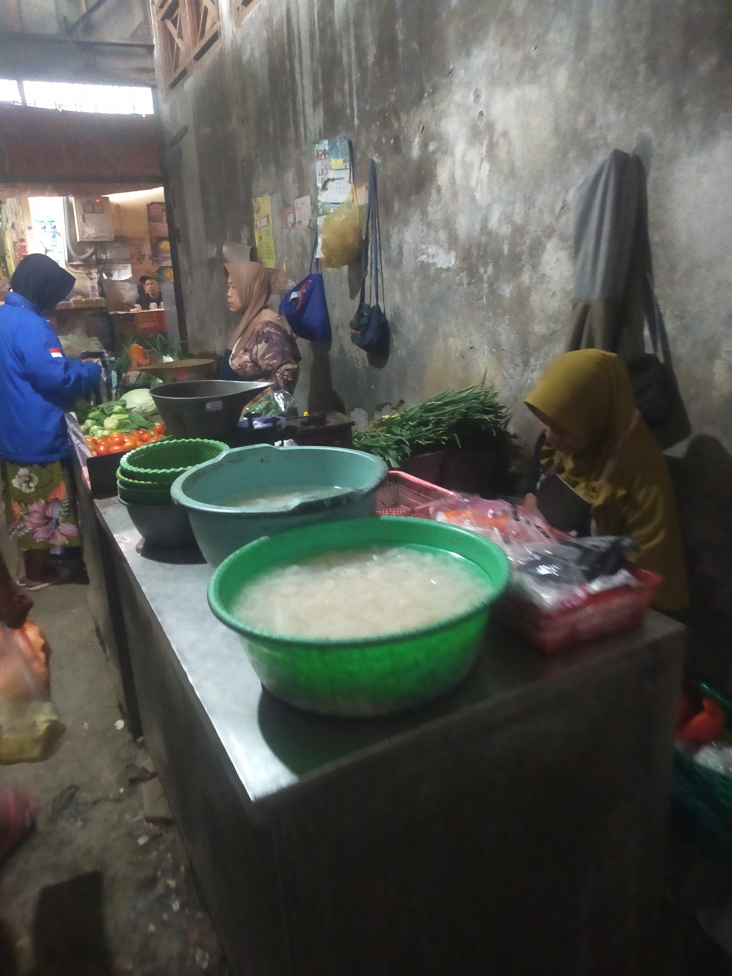 The image depicts a market scene in Trayeman, Indonesia. In the foreground, three large plastic bowls are visible on a stainless steel counter. The bowl on the left is green and appears empty, while the middle bowl is teal and contains a pale liquid. The bowl on the right is also green and filled with the same pale liquid. Behind these bowls, a variety of vegetables, including tomatoes and leafy greens, are displayed.
To the left, a person wearing a blue jacket and a black headscarf is interacting with the vegetables. Further back, another person in a light brown headscarf and patterned clothing is standing near a large black basin. To the right, a person wearing a yellow headscarf is seated, preparing food with a knife. They are surrounded by baskets and plastic packaging containing items that are difficult to discern due to the image quality.
The environment appears to be an indoor market stall with rough concrete walls and wooden ceiling beams. Bags and other items hang on the wall in the background. The lighting is dim, suggesting it may be early morning or late afternoon. The ground is concrete and appears slightly dirty.